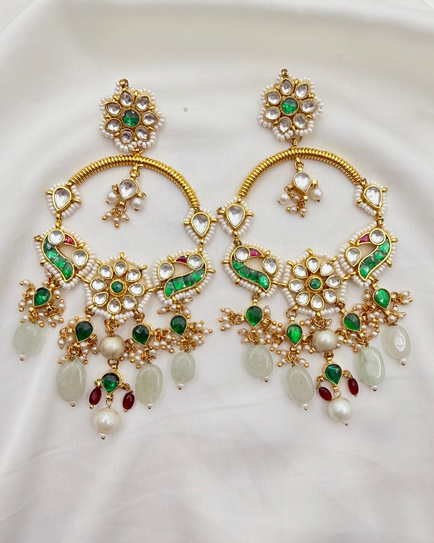A pair of ornate gold-toned traditional Indian chandelier earrings decorated with green and red enamel, white faux pearls, and light green stone drops, displayed on a white fabric background.