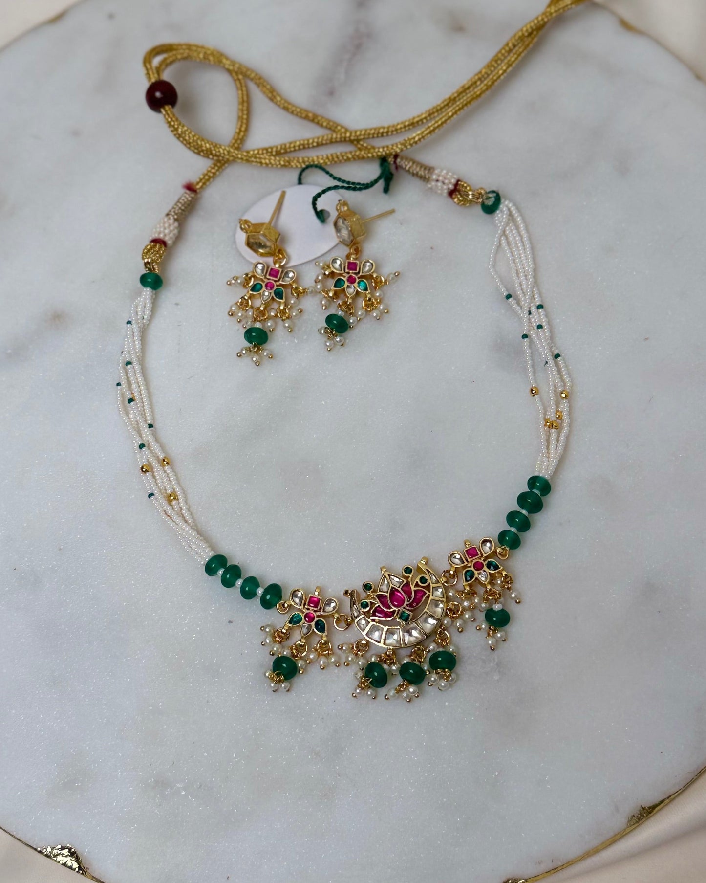 A traditional Indian jewelry set featuring a necklace, earrings, and a headpiece, displayed on a round white marble slab. The jewelry is made of gold-plated metal with white beads, green stones, and red gemstone accents.