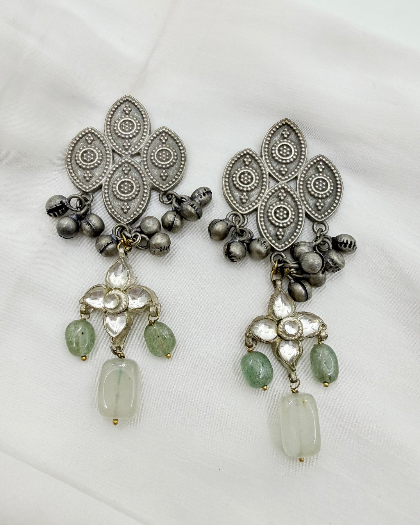 A pair of ornate silver-toned dangle earrings with intricate filigree patterns, small metallic bells, and light green stone accents, displayed on a white fabric background.