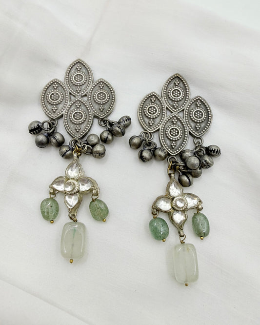 A pair of ornate silver-toned dangle earrings with intricate filigree patterns, small metallic bells, and light green stone accents, displayed on a white fabric background.