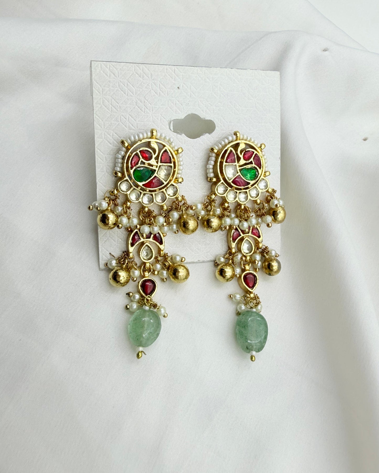 A pair of ornate gold-toned Indian jhumka earrings with green and red enamel details, white faux pearl beadwork, and green stone drops, displayed on a white card against a white fabric background.