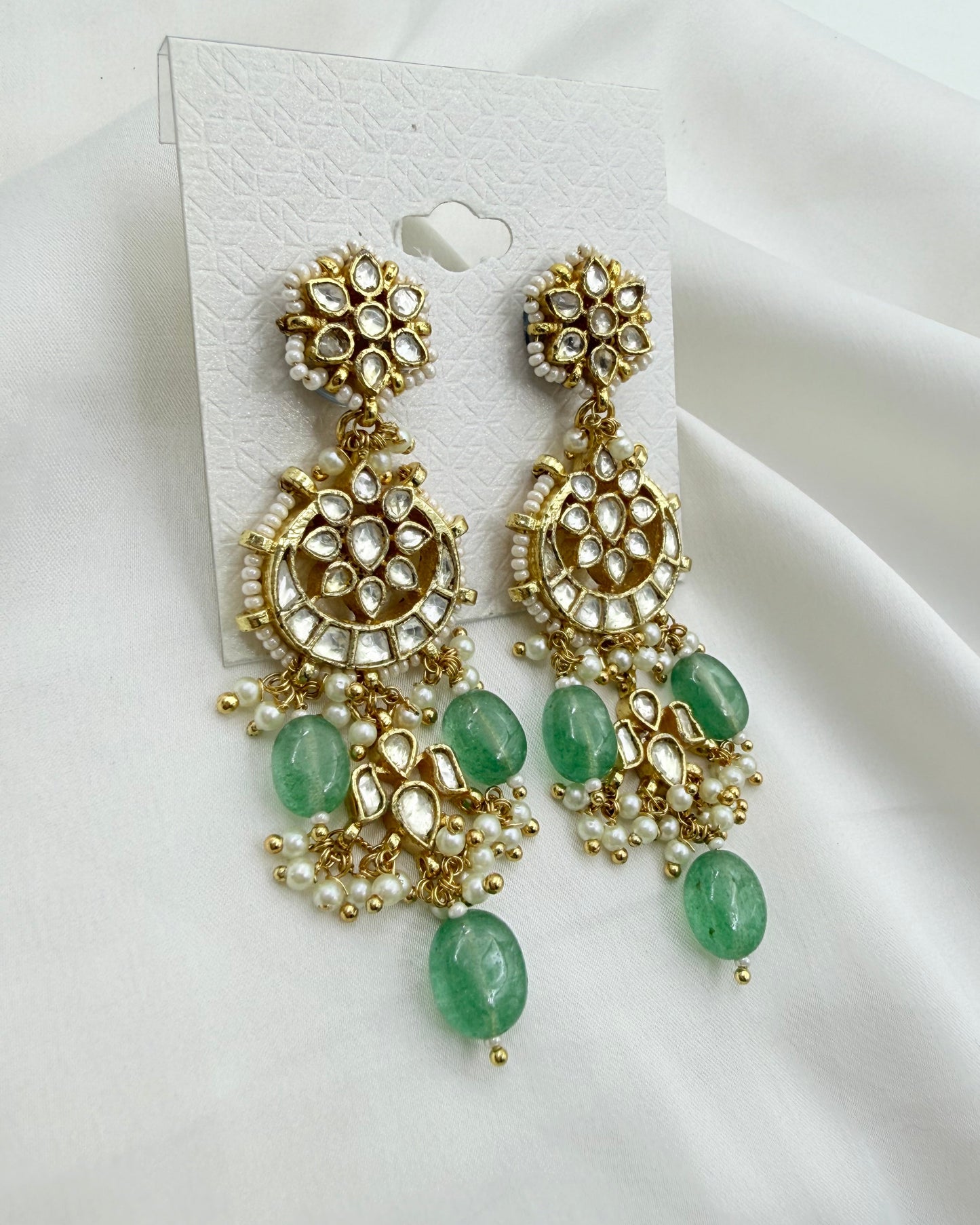 A pair of ornate gold-toned chandelier earrings with green stone drops and white pearl-like accents, displayed on a white card against a white fabric background.