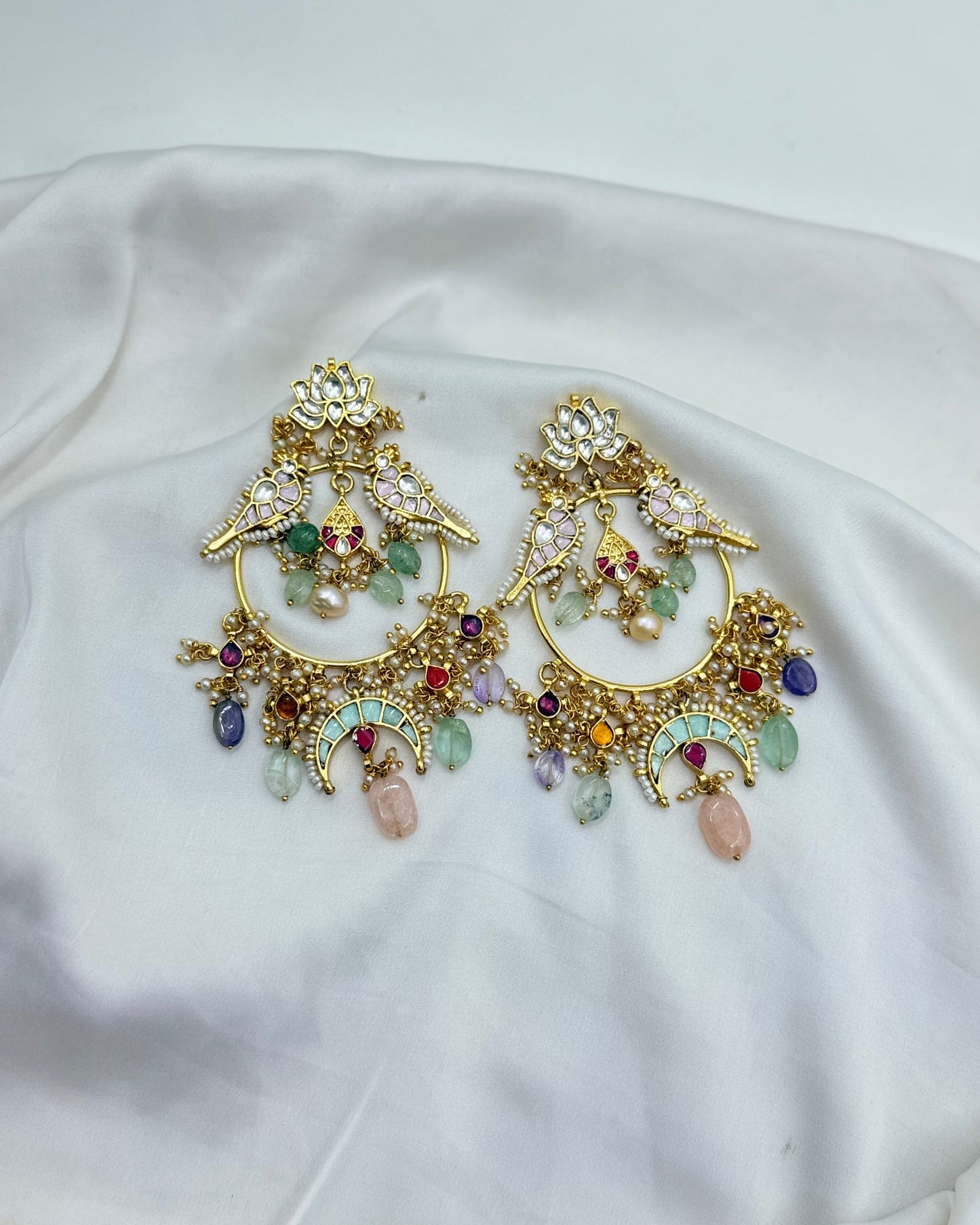 A pair of ornate gold-toned chandelier earrings decorated with multicolored gemstones, resting on draped white satin fabric.