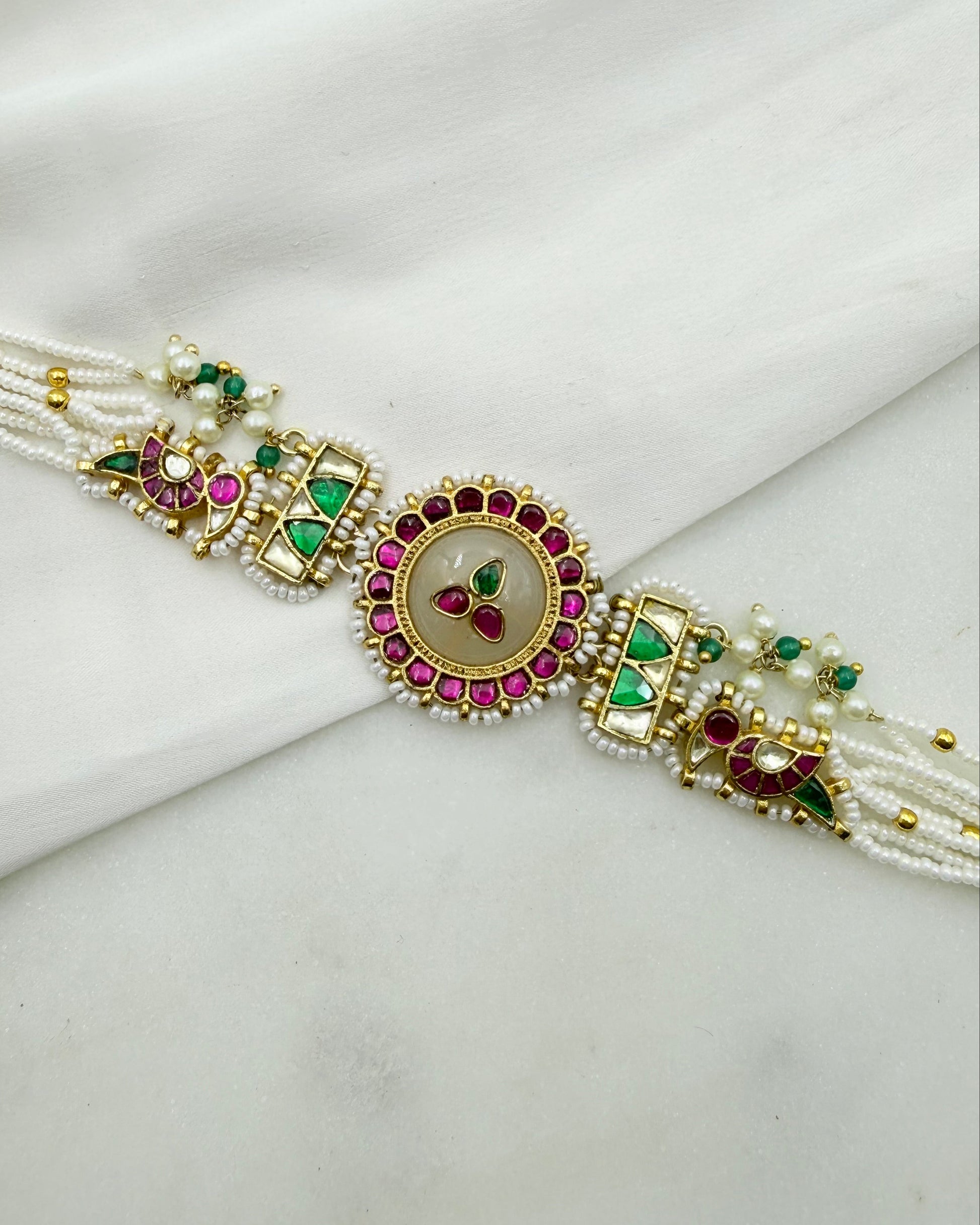 A traditional Indian Rakhi bracelet with a central oval pendant featuring a beige stone and pink and green stones, flanked by gold-plated motifs with green and pink accents, and white pearl strands on a white fabric background.