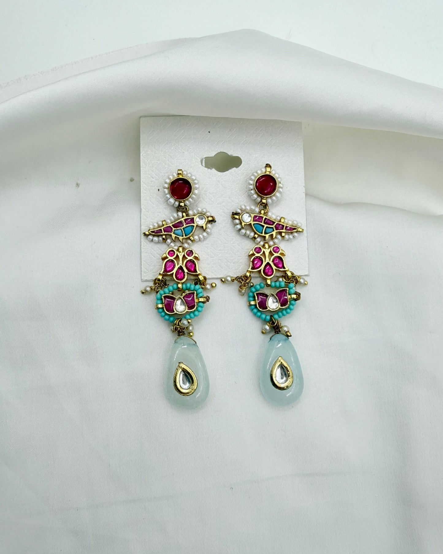 A pair of ornate gold-tone dangle earrings with red, pink, and turquoise stones, featuring light blue teardrop-shaped drops, displayed on a white card against a white fabric background.