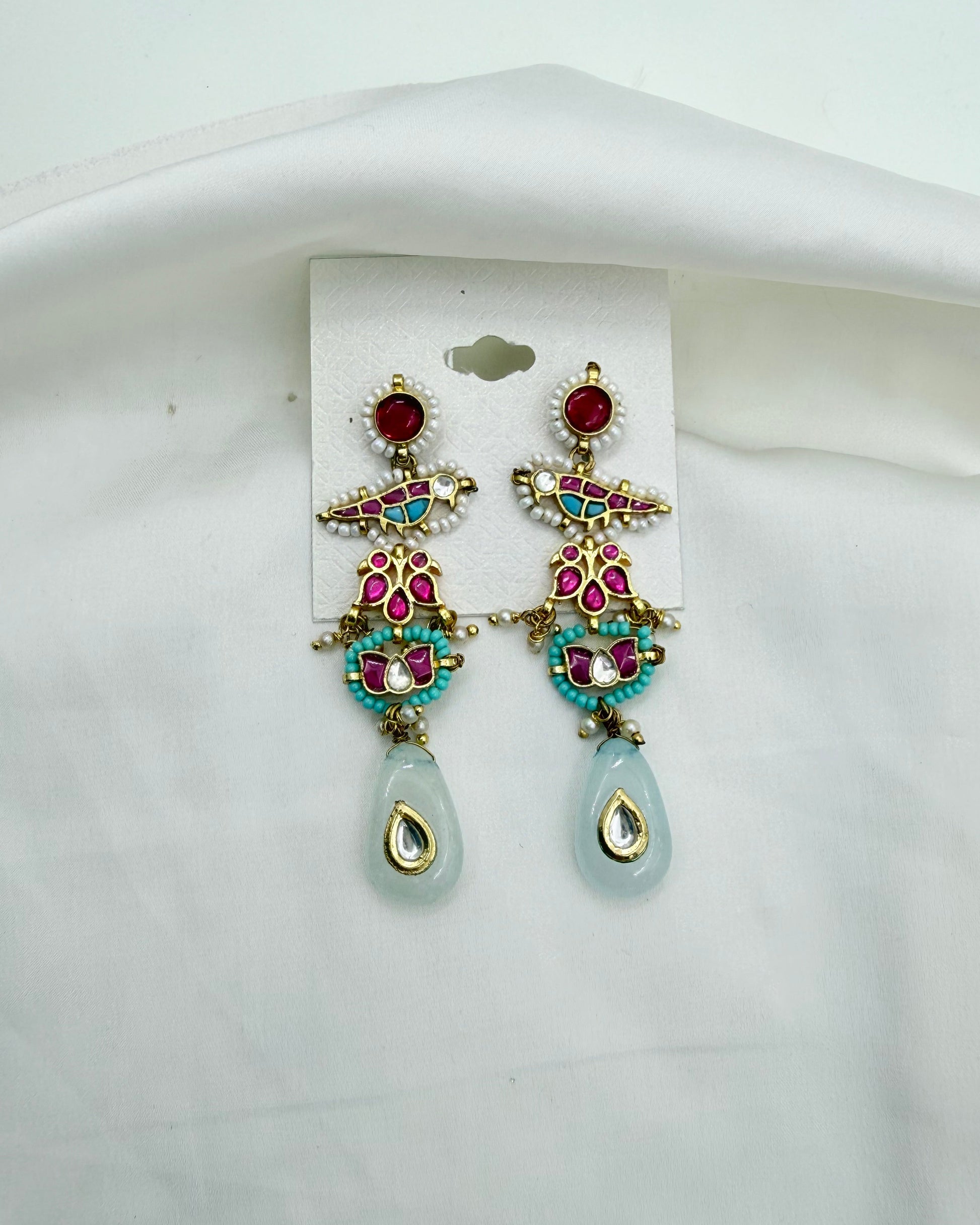 A pair of ornate gold-tone dangle earrings with red, pink, and turquoise stones, featuring light blue teardrop-shaped drops, displayed on a white card against a white fabric background.