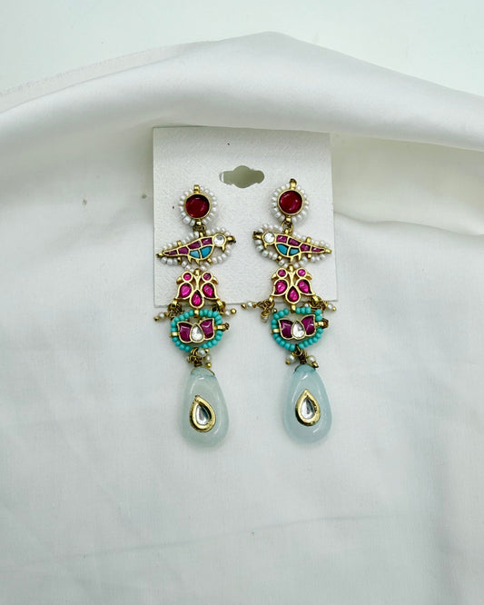 A pair of ornate gold-tone dangle earrings with red, pink, and turquoise stones, featuring light blue teardrop-shaped drops, displayed on a white card against a white fabric background.