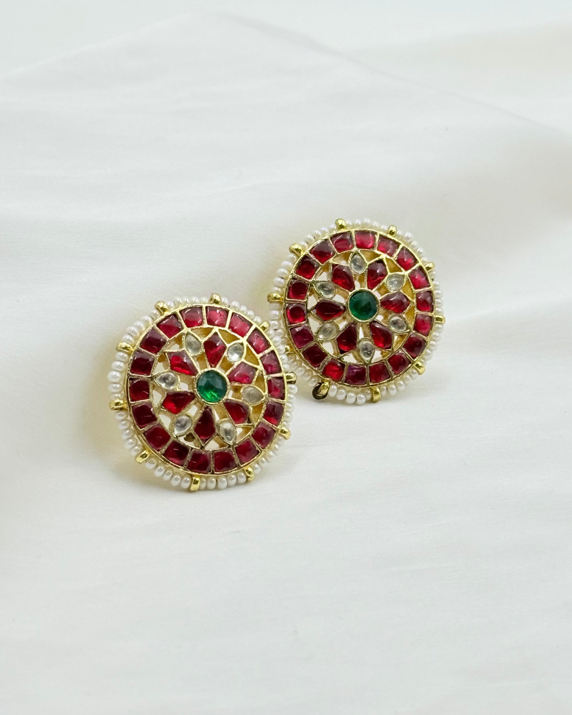 A pair of ornate circular gold-toned earrings decorated with red stones, white stones, and green center stones, displayed on a white fabric background.