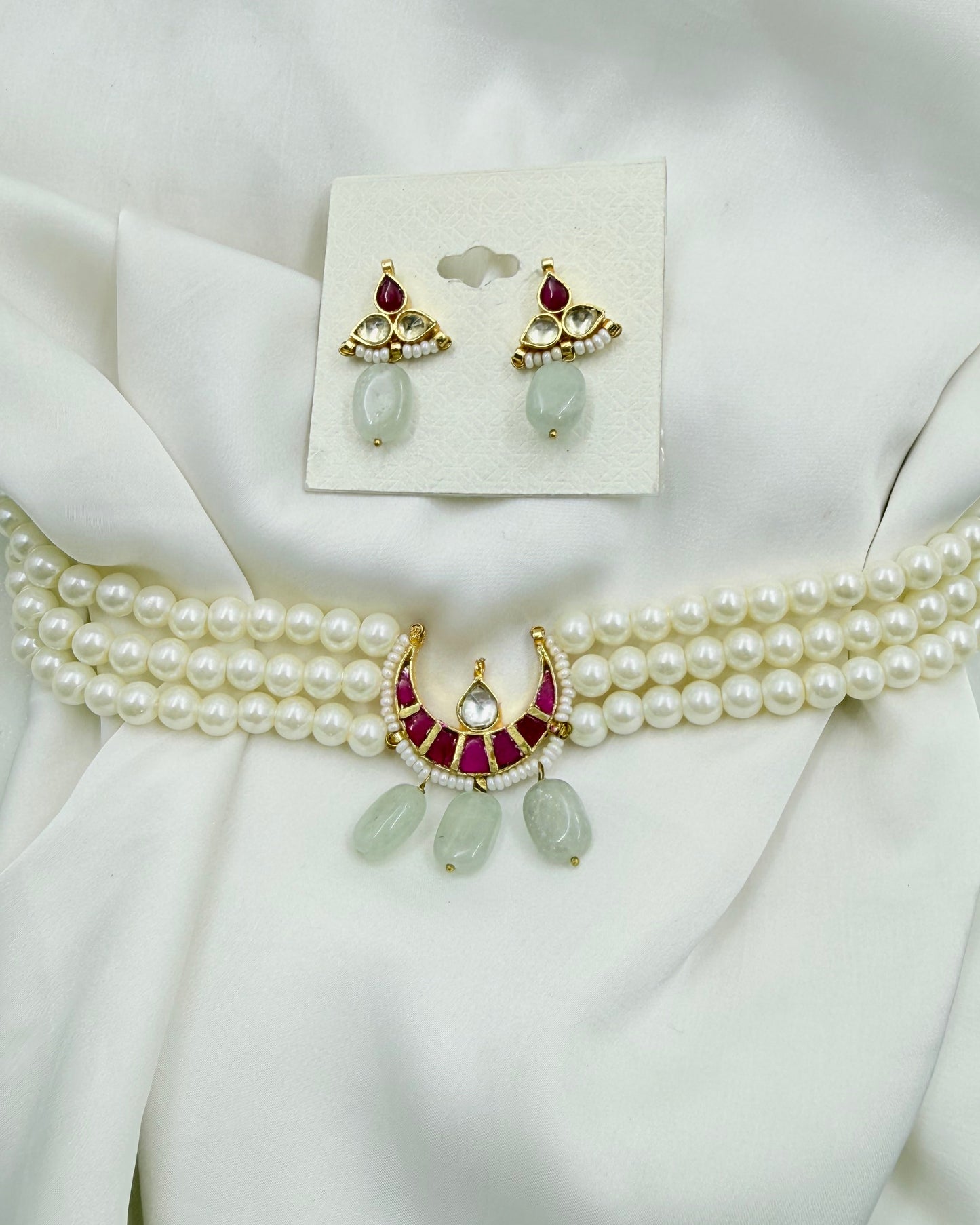 A gold and white pearl jewelry set featuring a multi-strand pearl choker with a crescent-shaped gold pendant set with red stones and light green teardrop beads, alongside matching gold and pearl earrings on a white backing card, all displayed on draped white satin fabric.