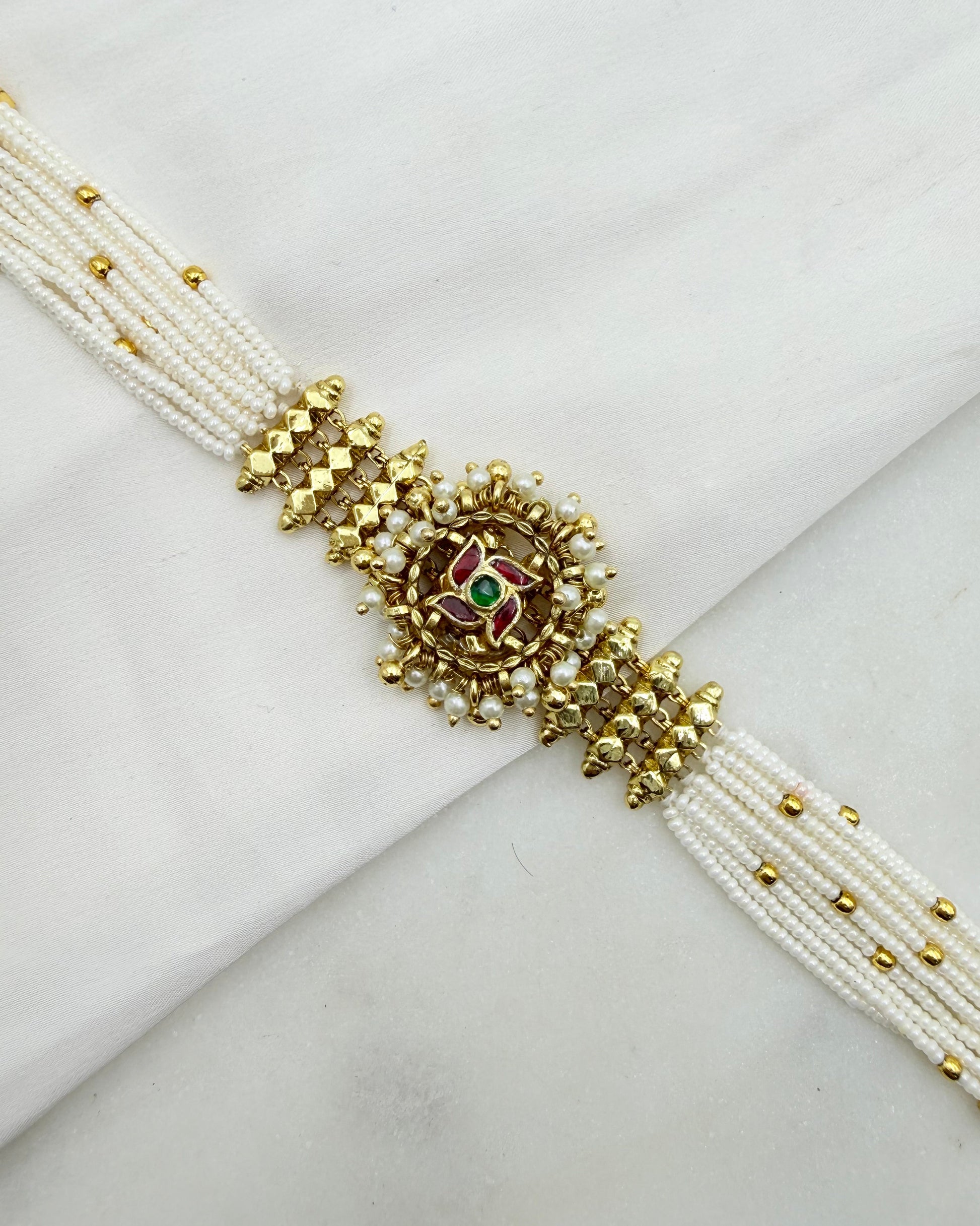 A traditional Indian multi-strand beaded bracelet, also known as a Kara, featuring white pearl-like beads, gold-plated metal borders, and a central decorative charm with gold, red, and green enamel details.