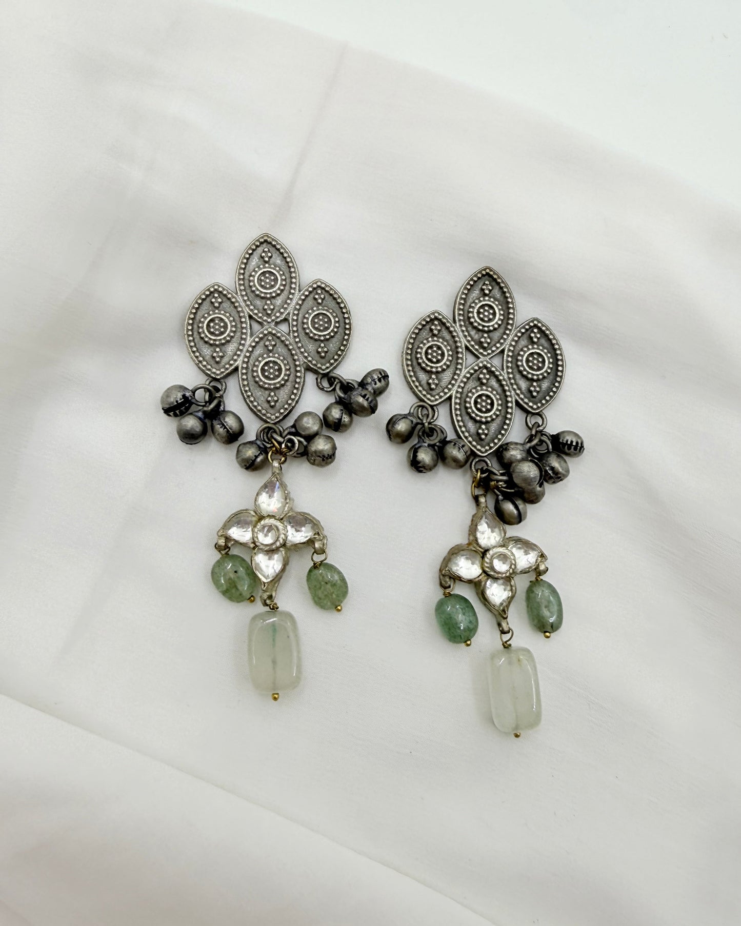A pair of ornate silver-toned traditional earrings with intricate filigree patterns, dark metallic beads, and green stone accents, displayed on a white fabric background.