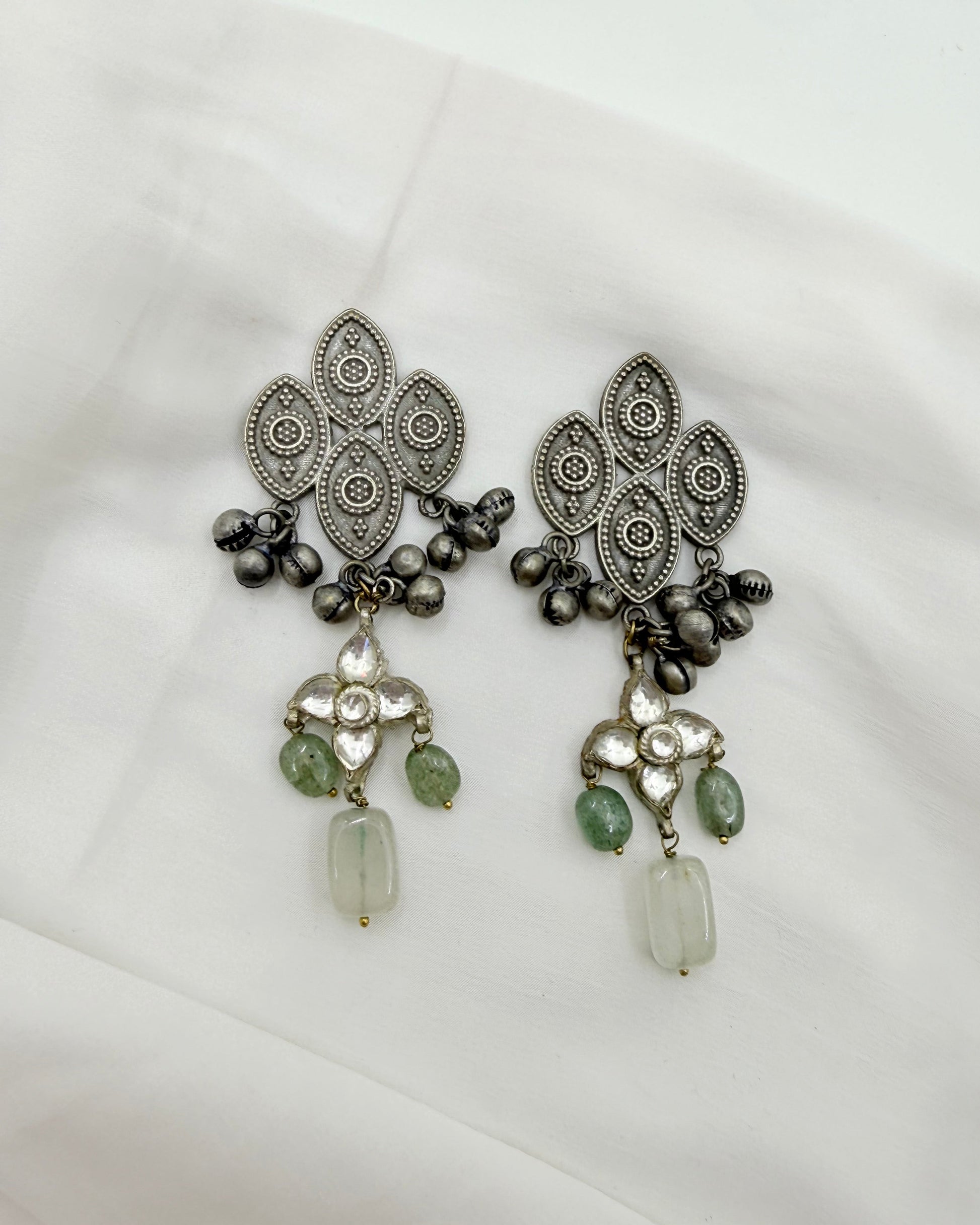 A pair of ornate silver-toned traditional earrings with intricate filigree patterns, dark metallic beads, and green stone accents, displayed on a white fabric background.