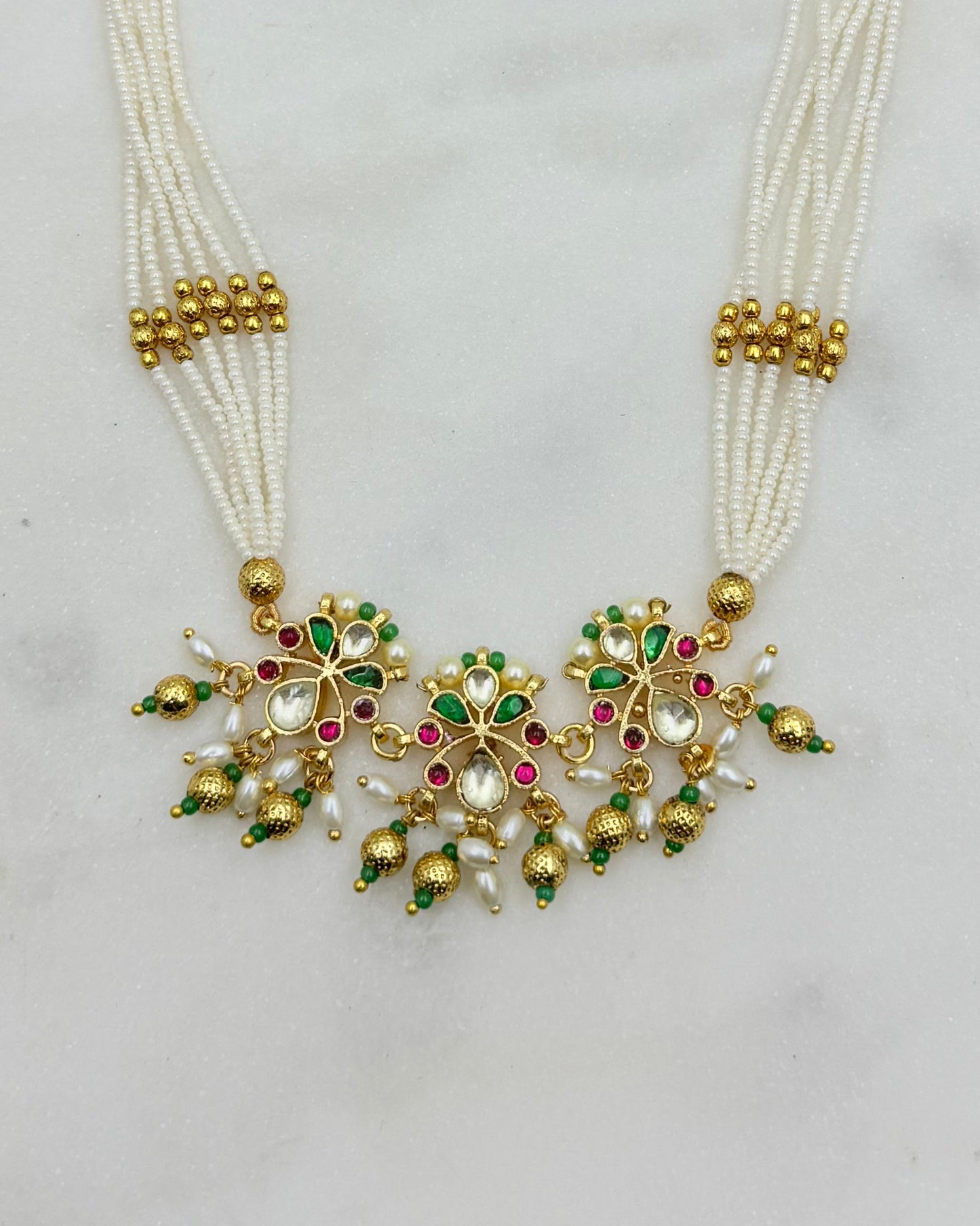 A traditional Indian multi-strand pearl necklace with a gold-plated pendant decorated with green and pink stones, set against a light gray background.
