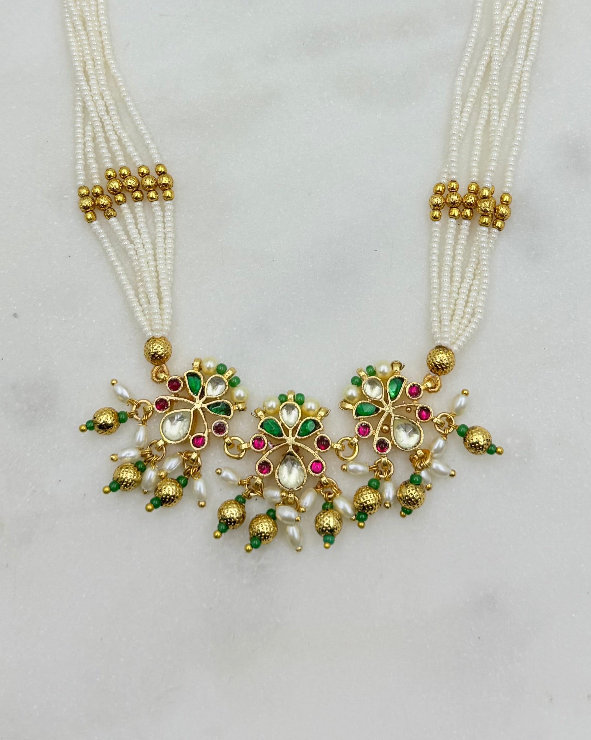 A traditional Indian multi-strand pearl necklace with a gold-plated pendant decorated with green and pink stones, set against a light gray background.