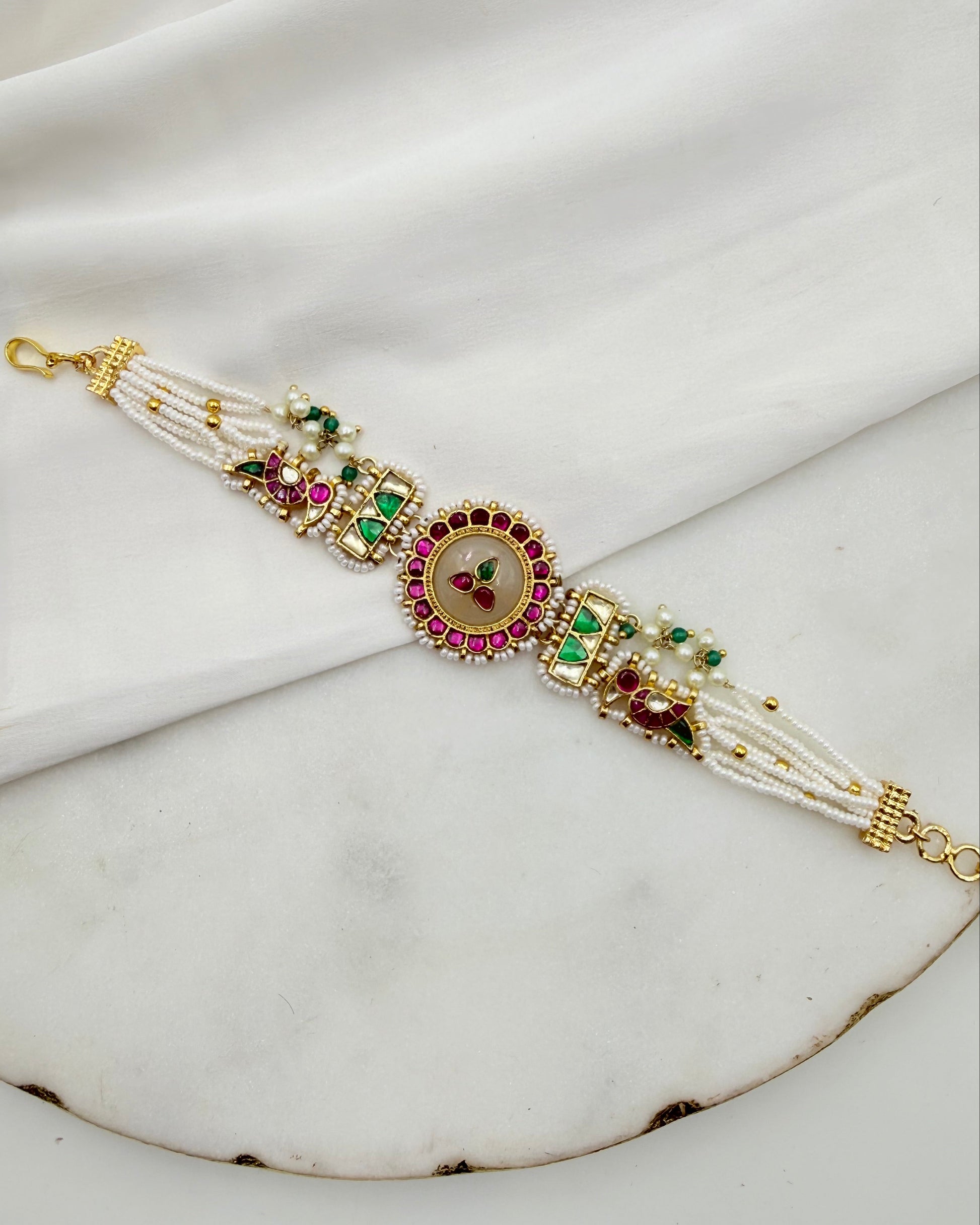 A traditional Indian gold-plated bracelet (Kara) decorated with white pearl strands, green and pink stones, and a central floral motif, displayed on a draped white fabric background.