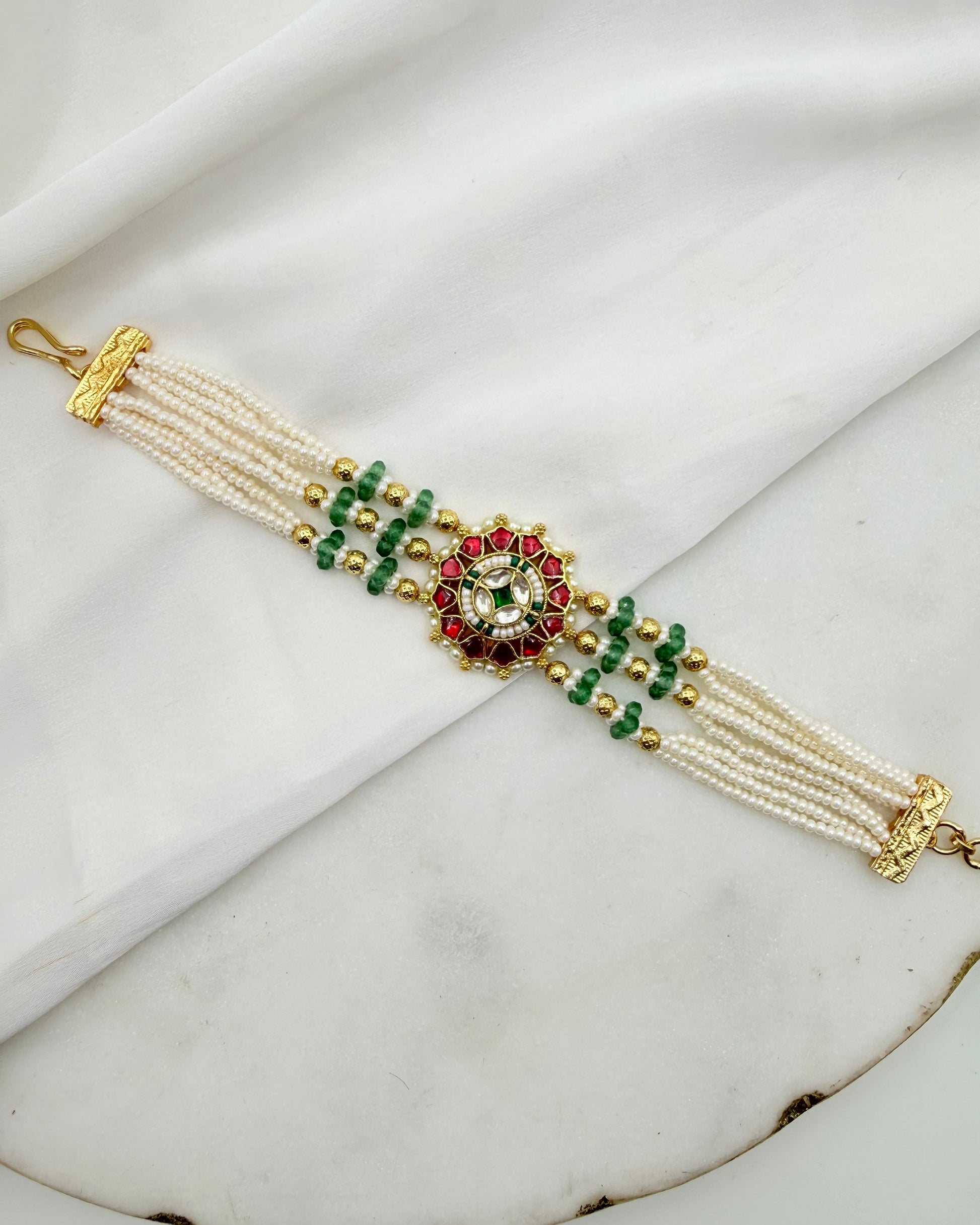 A multi-strand pearl bracelet with gold-plated metal details, green and red beads, and a central floral motif, displayed on a white draped fabric background.