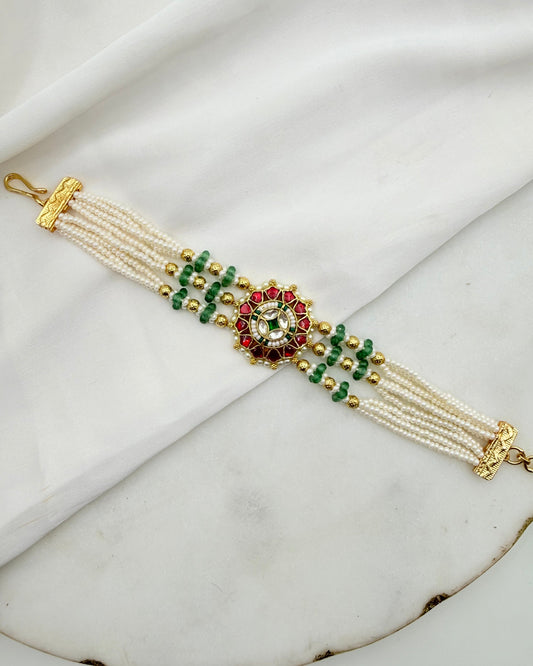 A multi-strand pearl bracelet with gold-plated metal details, green and red beads, and a central floral motif, displayed on a white draped fabric background.