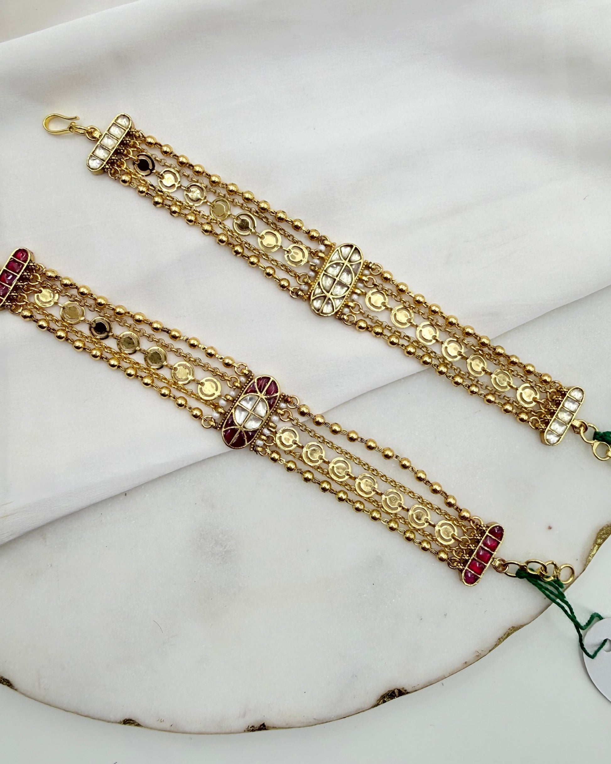 Two ornate gold-toned traditional bracelets with intricate beadwork, mirror work, and small red and white stone accents, displayed on a white fabric surface.