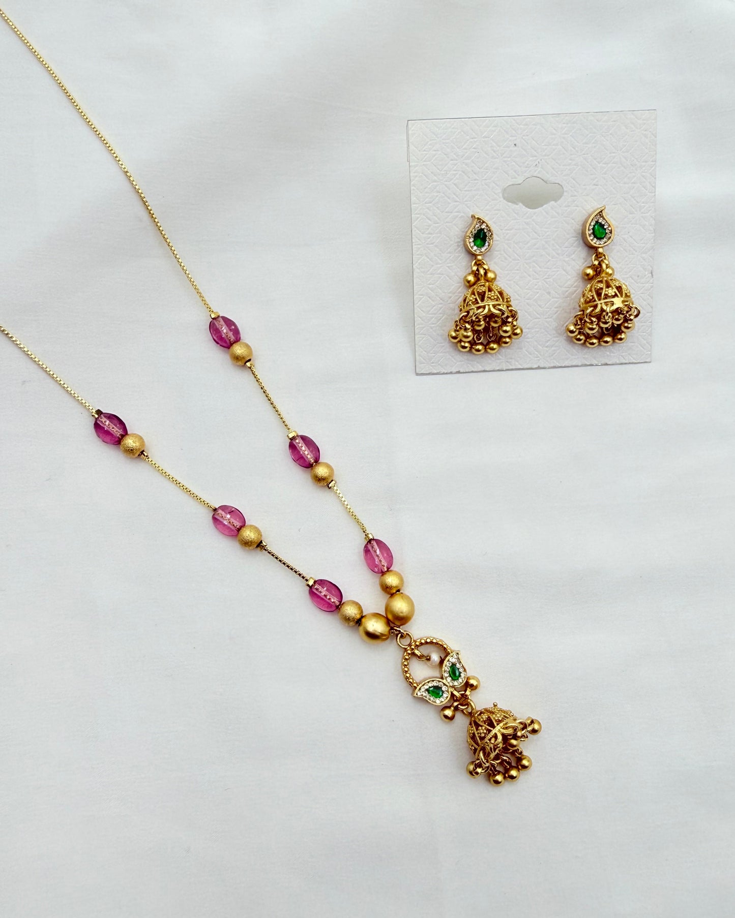 Gold-toned jewelry set featuring a necklace and matching earrings with pink faceted beads, gold spacer beads, and small green stone accents, displayed on a white surface with the earrings shown on a small card.