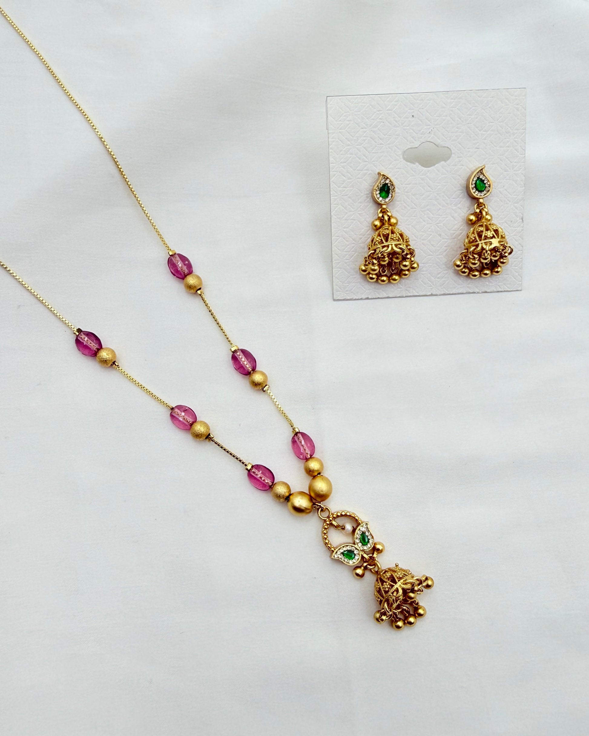 Gold-toned jewelry set featuring a necklace and matching earrings with pink faceted beads, gold spacer beads, and small green stone accents, displayed on a white surface with the earrings shown on a small card.