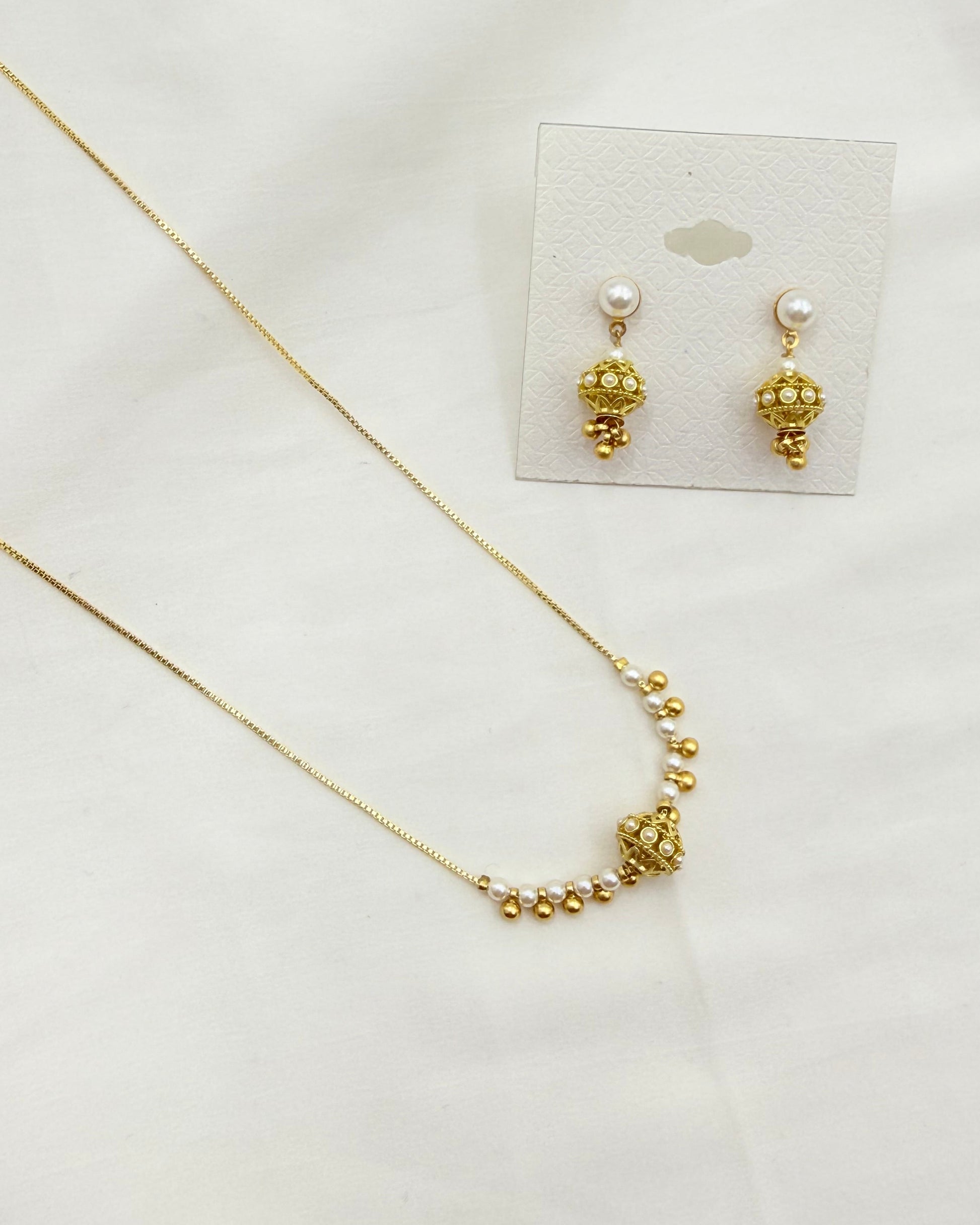 Gold jewelry set featuring a delicate chain necklace and matching stud earrings with pearl-like studs and textured gold drops, displayed on a white fabric background with a small card backing.