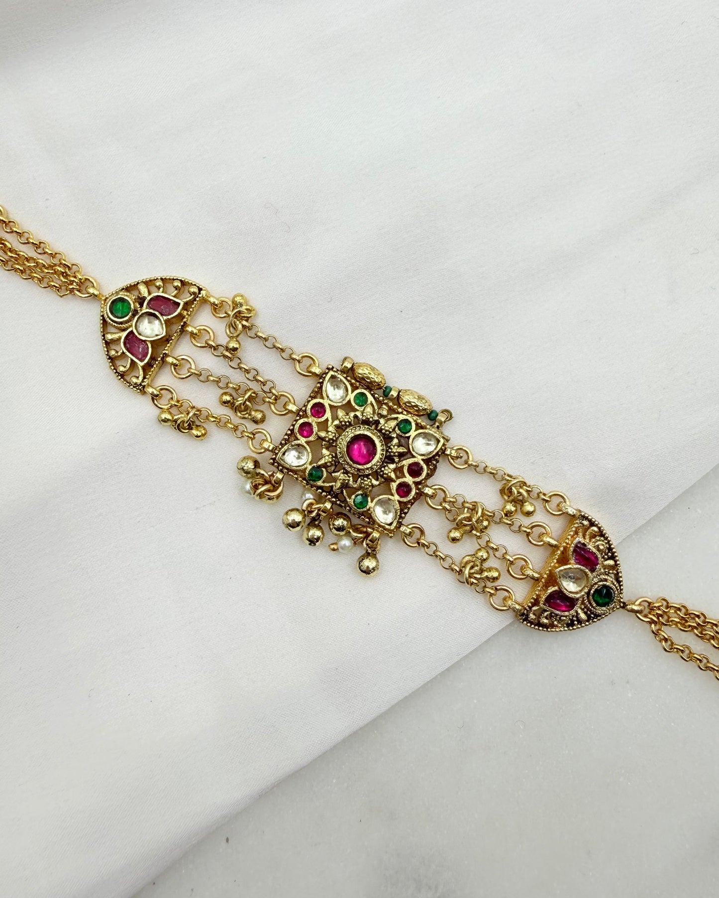Gold-toned traditional Indian jewelry piece featuring a central ornate pendant with red and green stones and dangling chain elements, displayed on a white fabric background.