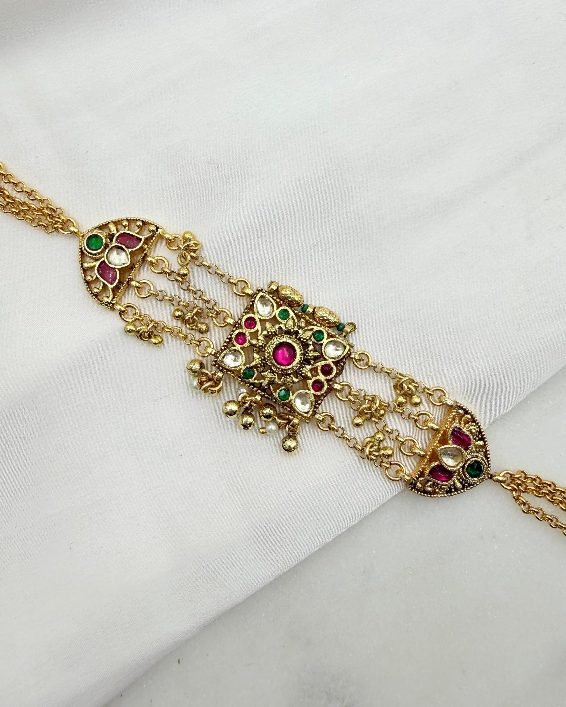 Gold-toned traditional Indian jewelry piece featuring a central ornate pendant with red and green stones and dangling chain elements, displayed on a white fabric background.