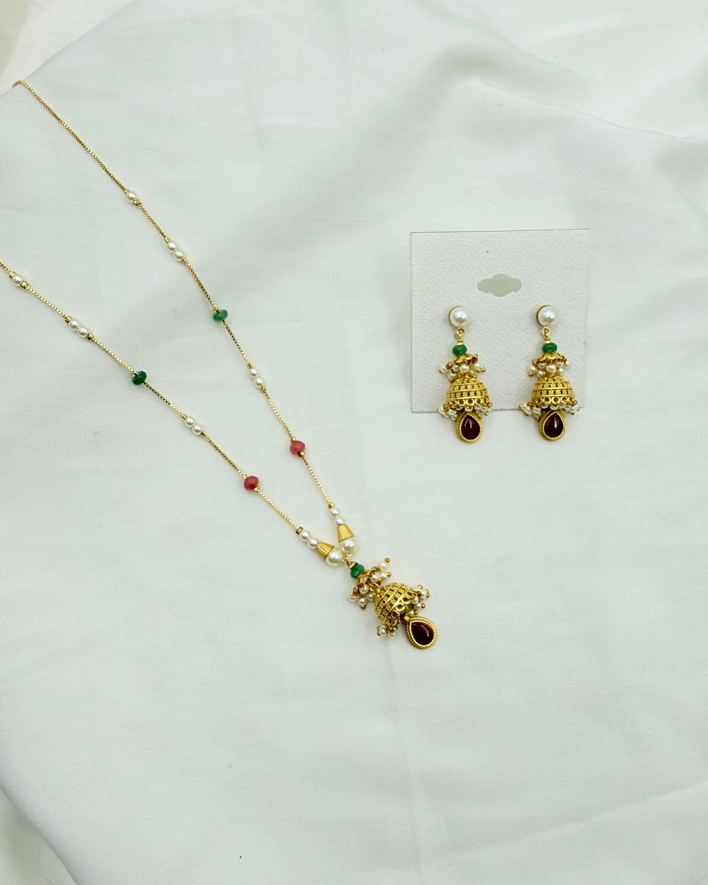 Gold-toned traditional jewelry set featuring a necklace and matching earrings with pearl-like white beads, green and red accent beads, and a decorative hanging pendant, displayed on a white fabric background.