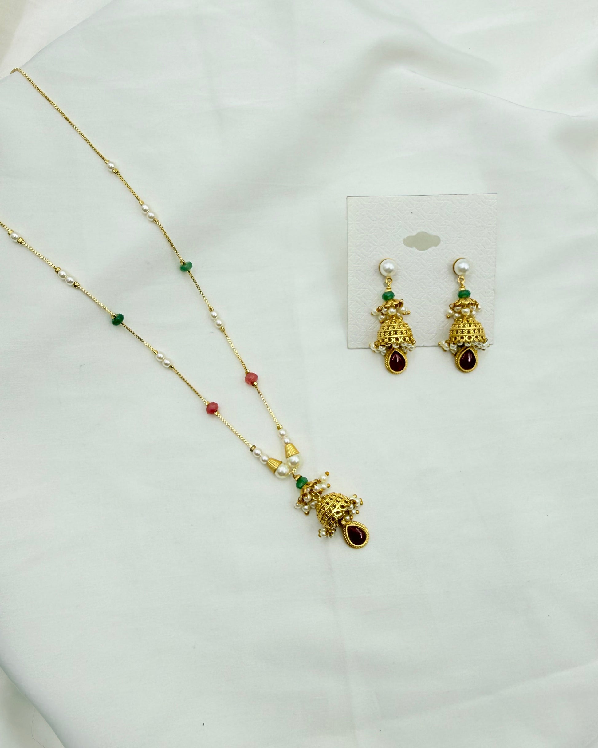 Gold-toned traditional jewelry set featuring a necklace and matching earrings with pearl-like white beads, green and red accent beads, and a decorative hanging pendant, displayed on a white fabric background.