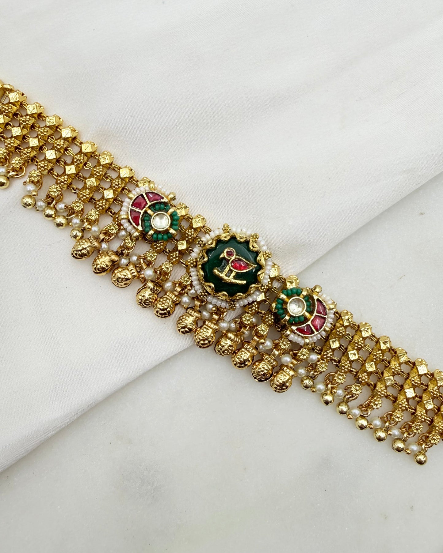 Gold-toned traditional Indian jewelry piece featuring intricate beadwork, small pearl-like beads, and three decorative floral motifs with green, red, and white enamel details, displayed on a white fabric background.