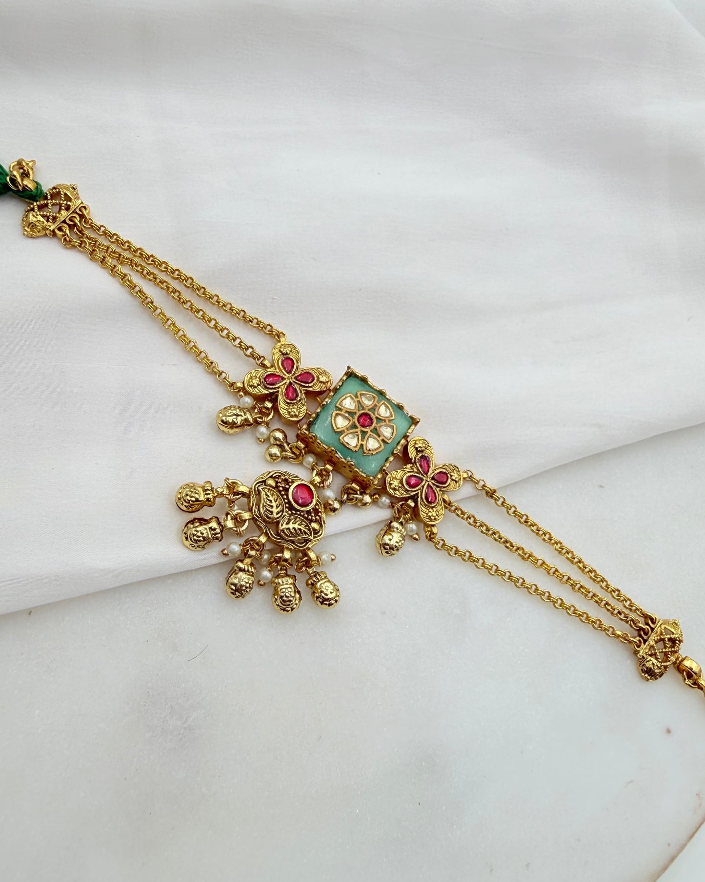 An ornate gold-toned traditional necklace with a rectangular turquoise green centerpiece, floral motifs, and small red stone accents, displayed on a white fabric background.