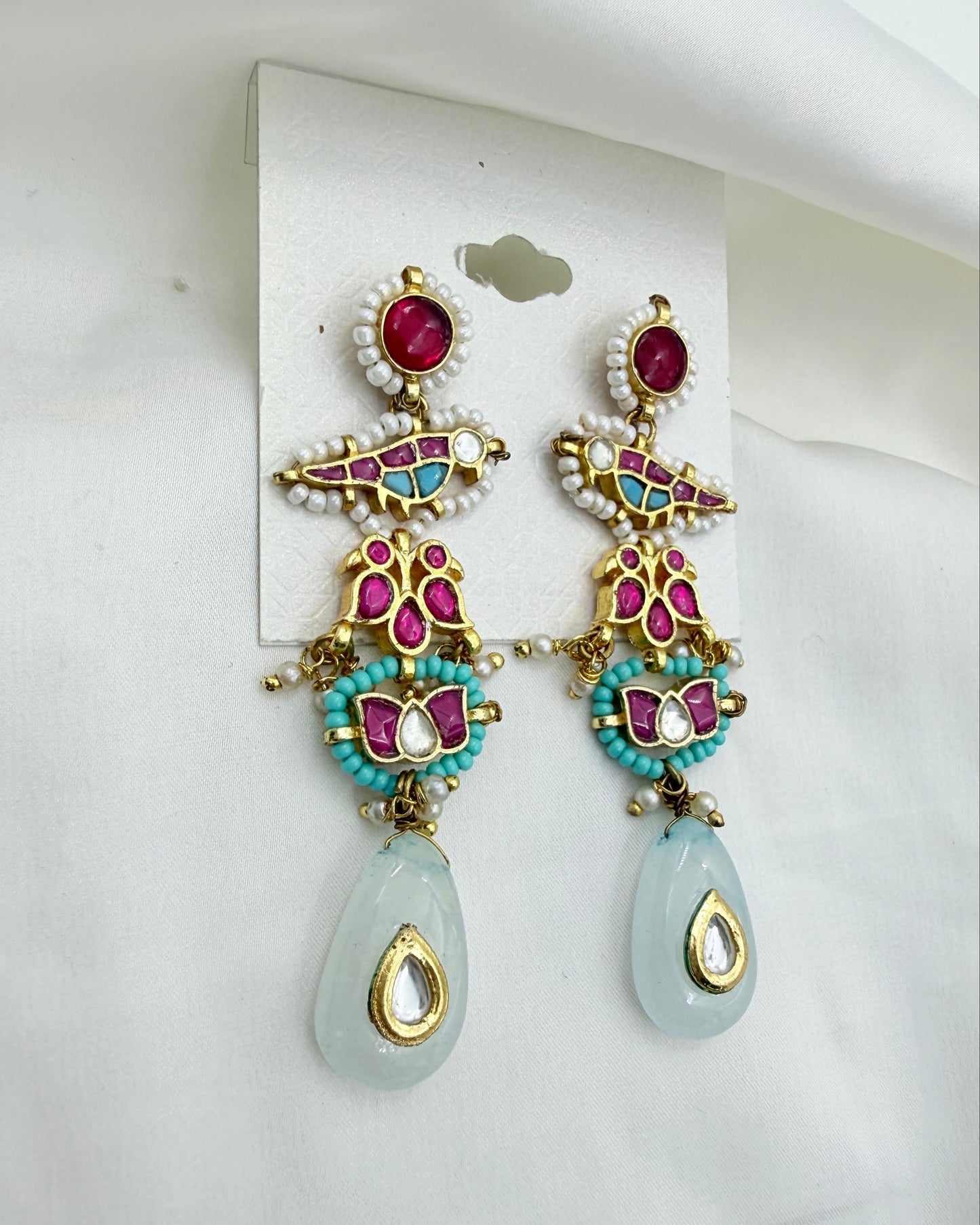 A pair of ornate gold-plated dangle earrings with turquoise, pink, and white beadwork, featuring a light blue-green teardrop stone at the bottom, displayed on a white card against a white fabric background.