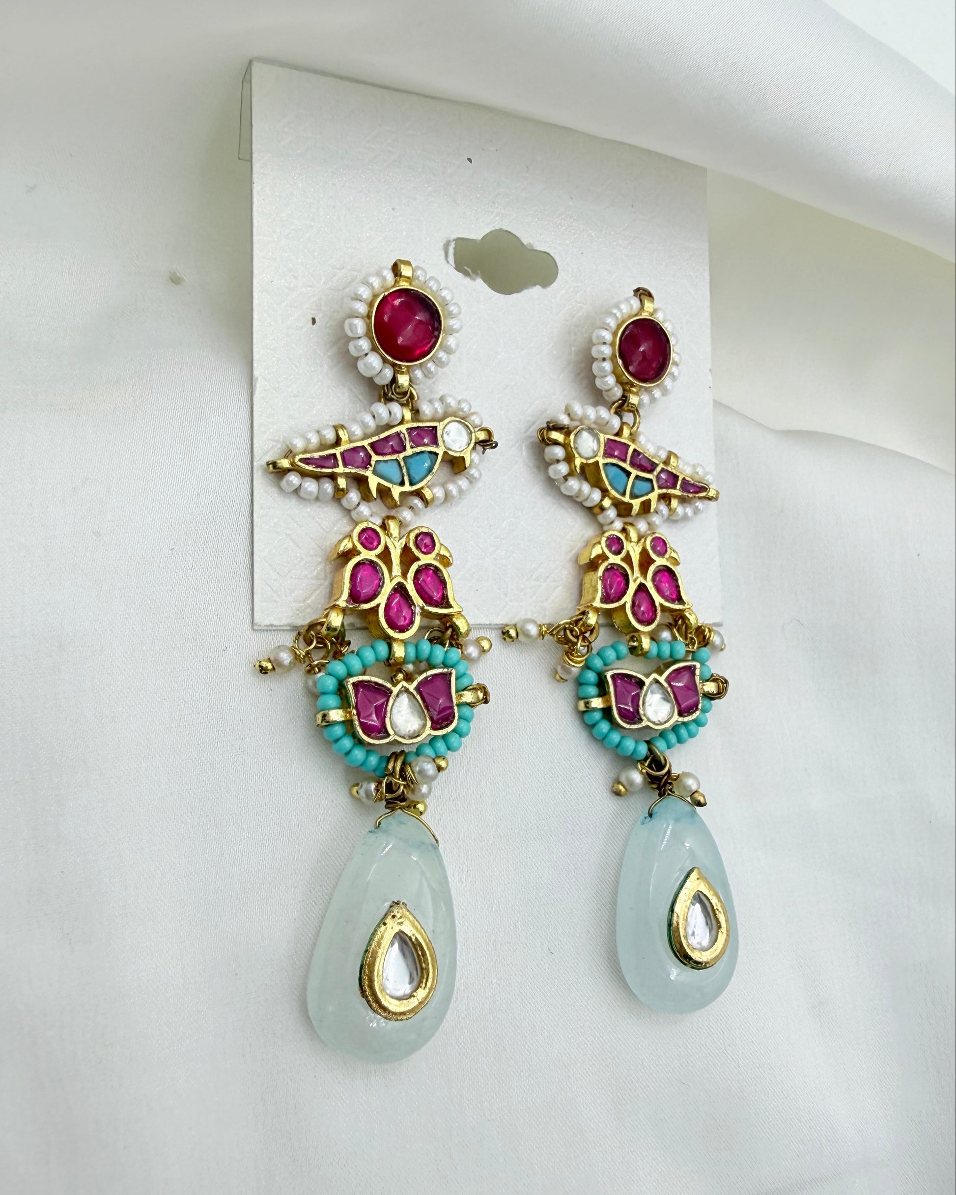 A pair of ornate gold-plated dangle earrings with turquoise, pink, and white beadwork, featuring a light blue-green teardrop stone at the bottom, displayed on a white card against a white fabric background.
