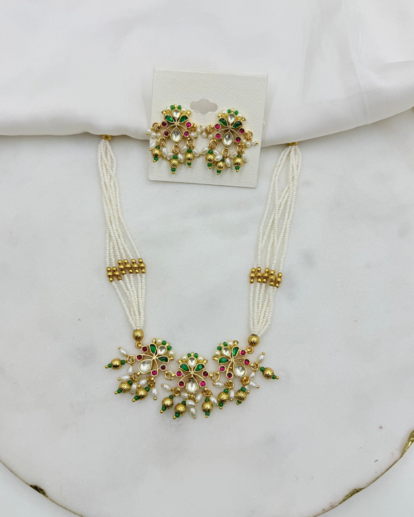 A traditional Indian jewelry set featuring a necklace and matching earrings. The necklace has multiple strands of white beads with a central gold-plated floral motif embellished with green, pink, and white stones.