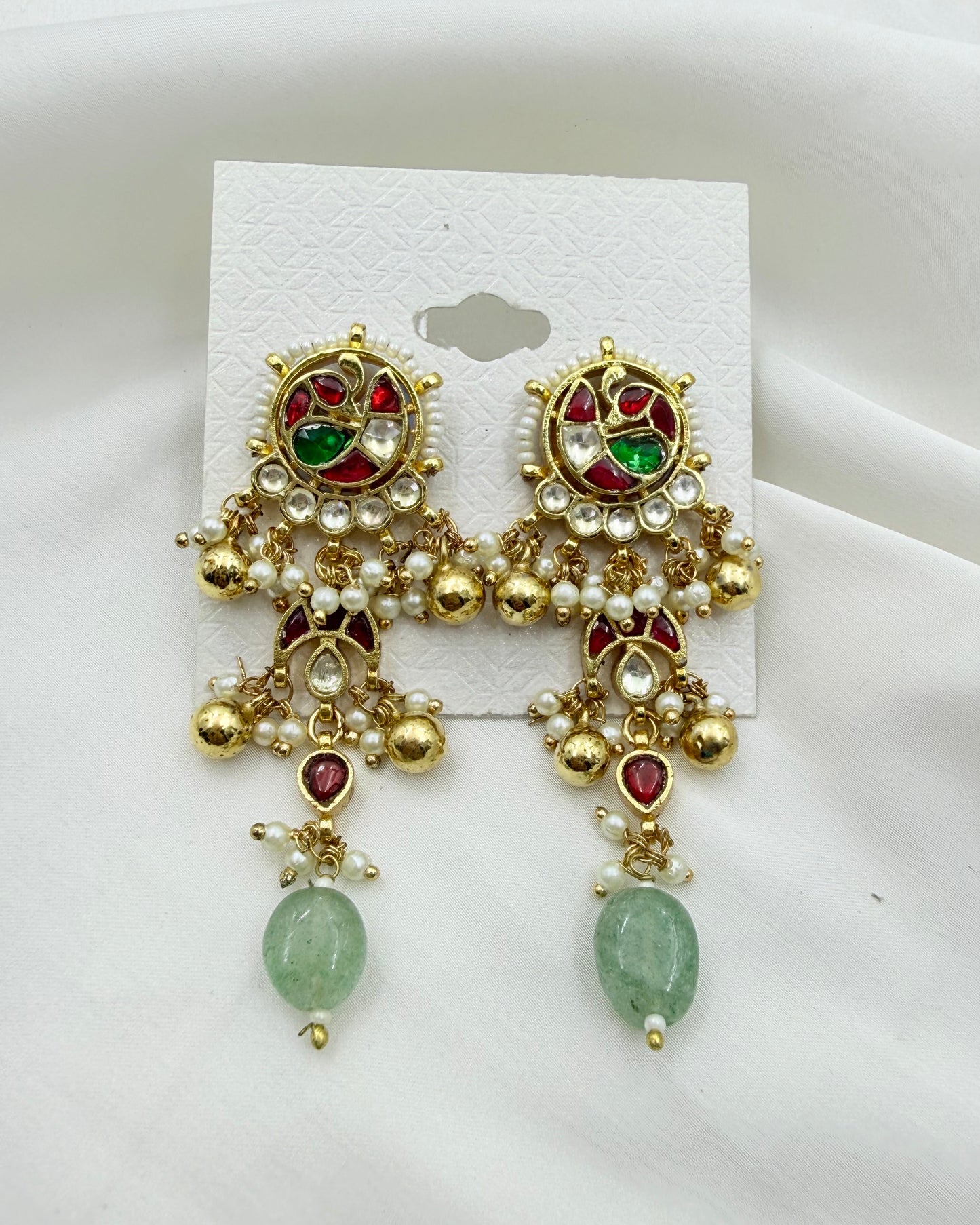 A pair of ornate gold-toned traditional Indian earrings with red and green stones, white faux pearls, and green bead drops, displayed on a white card against a white fabric background.