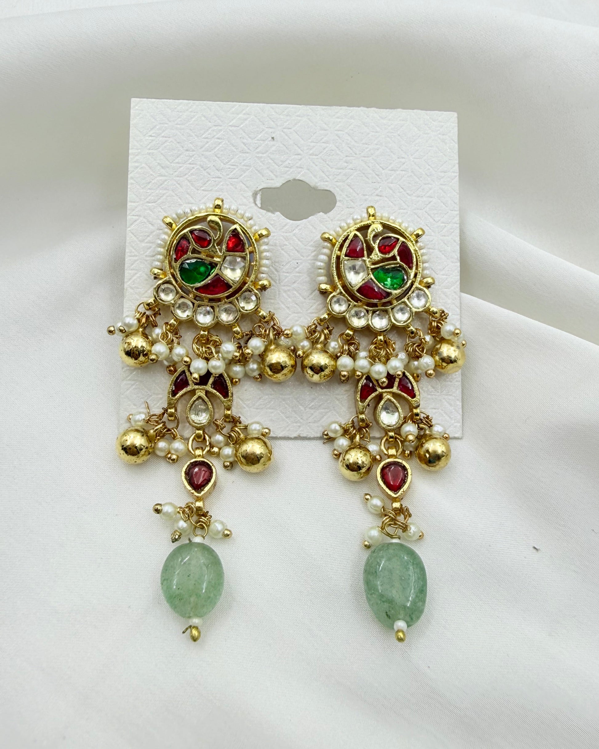 A pair of ornate gold-toned traditional Indian earrings with red and green stones, white faux pearls, and green bead drops, displayed on a white card against a white fabric background.