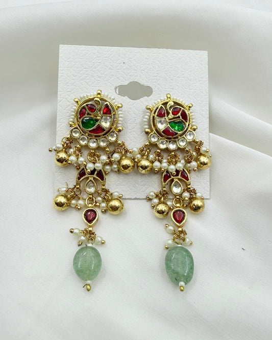 A pair of ornate gold-toned traditional Indian earrings with red and green stones, white faux pearls, and green bead drops, displayed on a white card against a white fabric background.