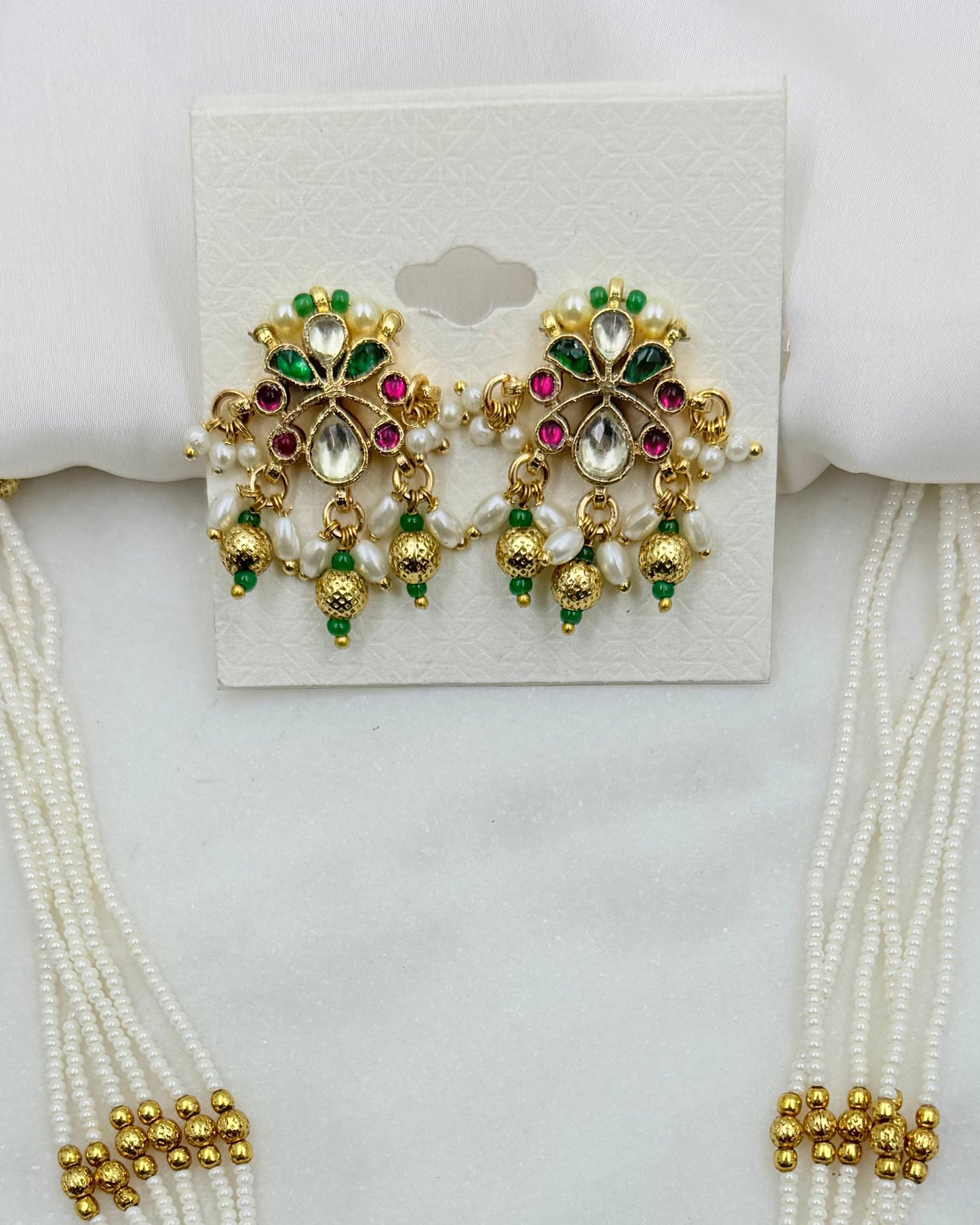 Gold-plated Indian bridal jewelry set featuring a pair of floral chandelier earrings with white pearls, green and pink stones, and gold beads, displayed on a white card with matching pearl strands in the background.