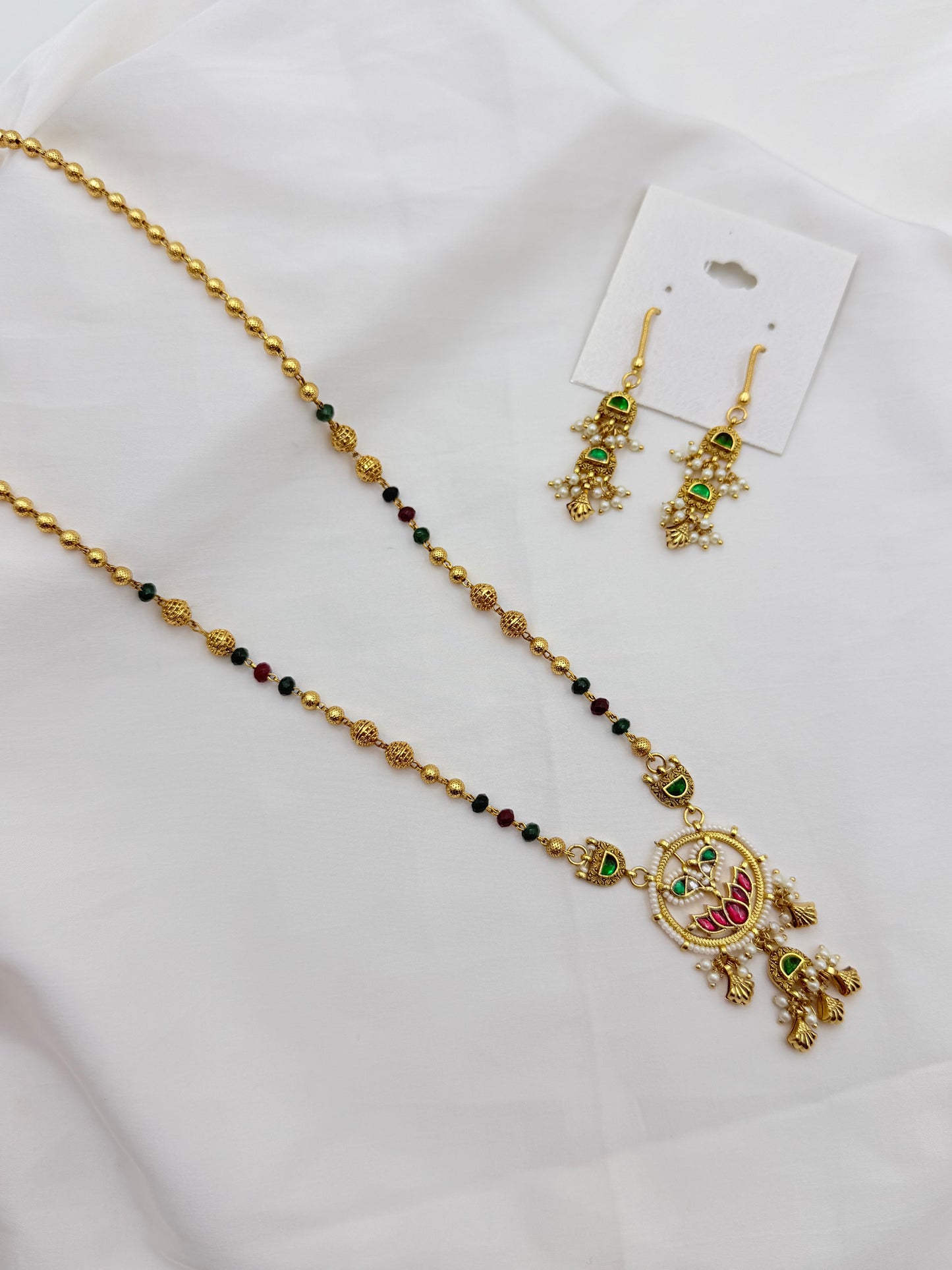 Gold-toned traditional necklace and matching drop earrings laid on a white fabric background. The necklace has small dark beads and a decorative pendant with green and red stone accents, and the earrings are attached to a white display card.