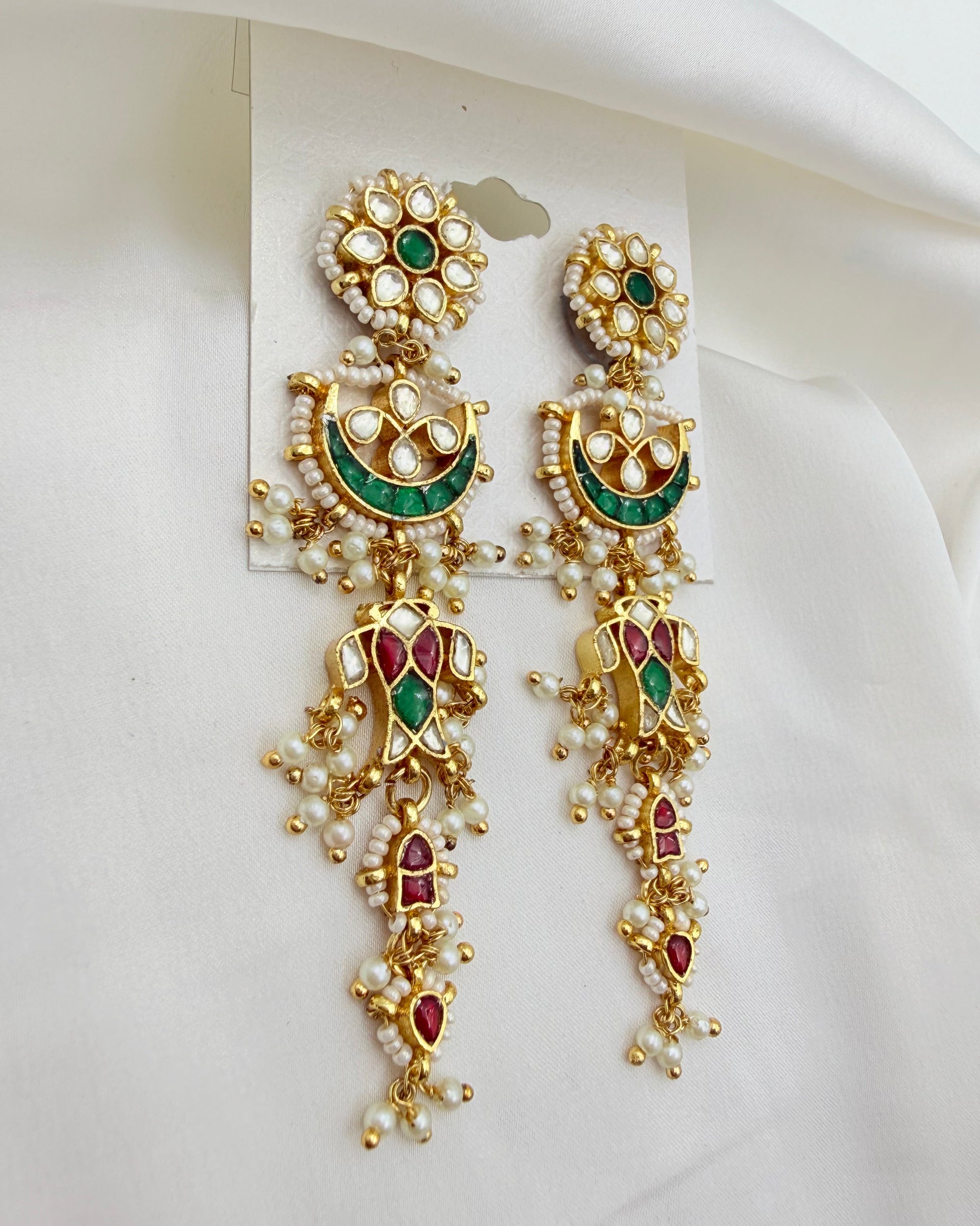 A pair of ornate gold-plated Indian Kundan dangle earrings decorated with white faux pearls, green enamel, and red stones, displayed on a white card against a white fabric background.