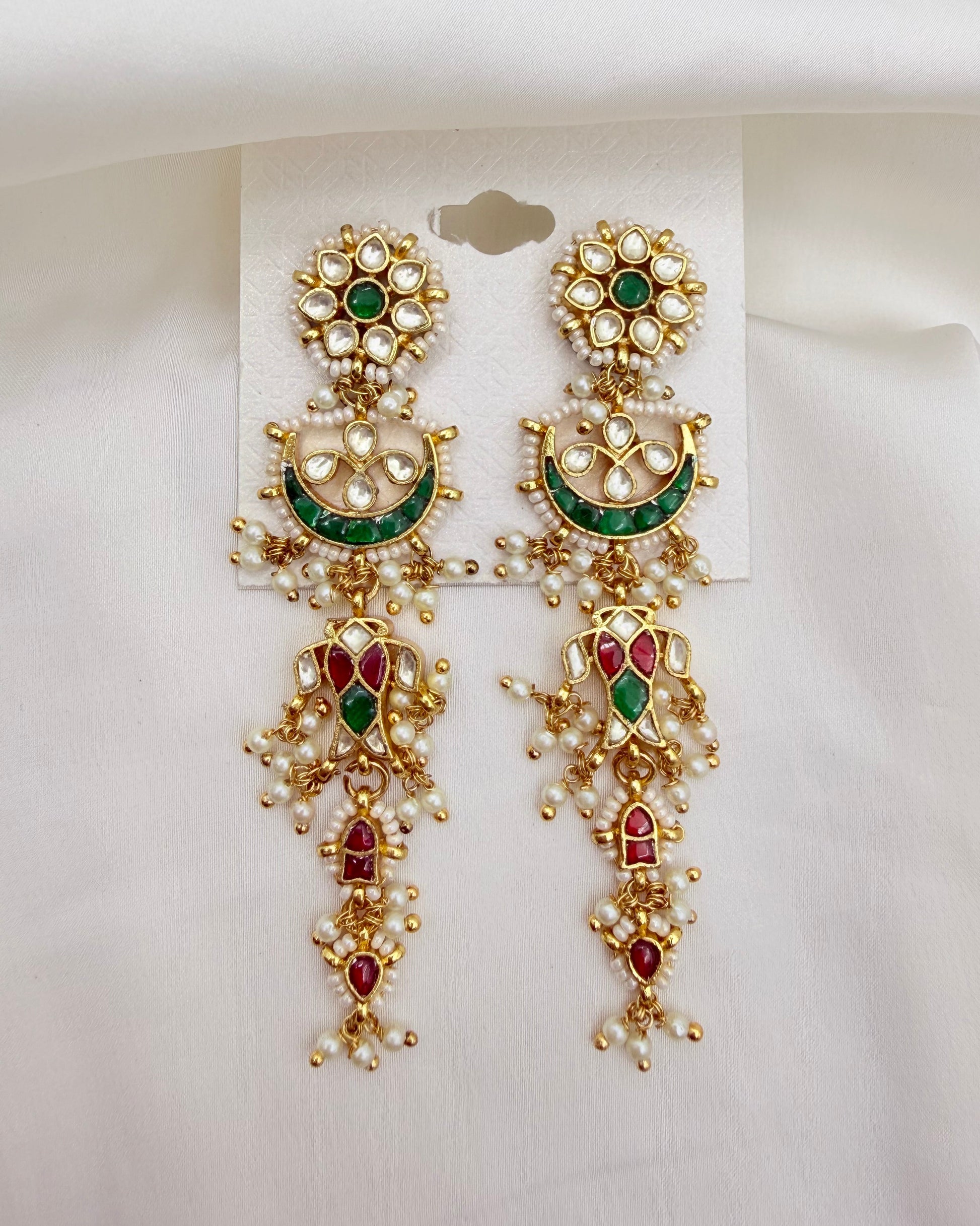 A pair of ornate gold-toned Indian chandelier earrings with green and red enamel details and white pearl-like stones, displayed on a white backing card against a white fabric background.