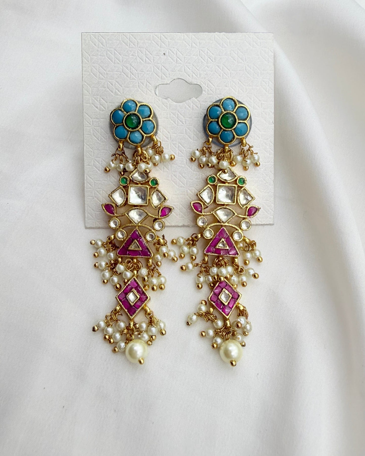 A pair of ornate gold-toned dangle earrings with turquoise blue floral studs, pink enamel details, and white faux pearls, displayed on a white card against a white fabric background.