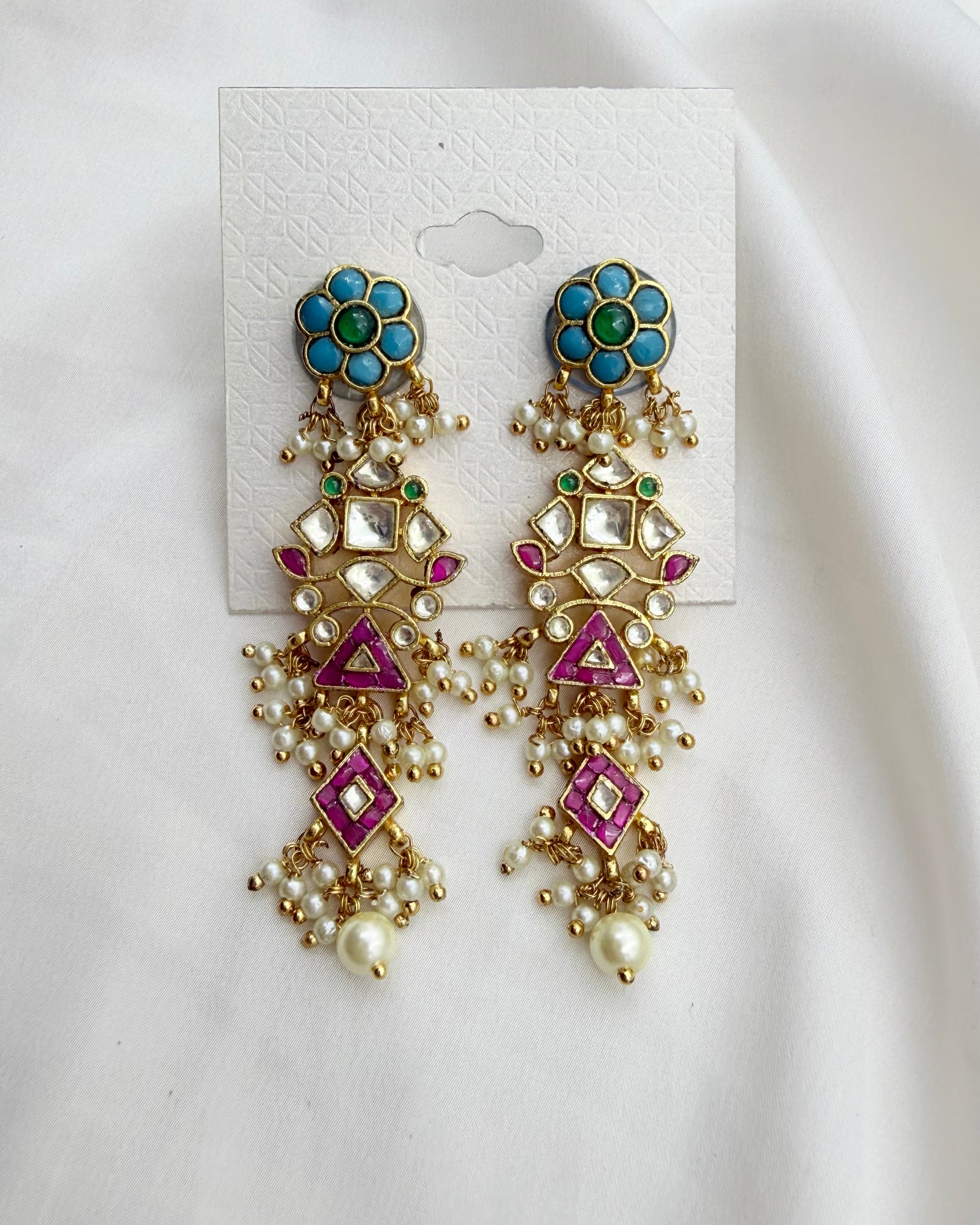 A pair of ornate gold-toned dangle earrings with turquoise blue floral studs, pink enamel details, and white faux pearls, displayed on a white card against a white fabric background.