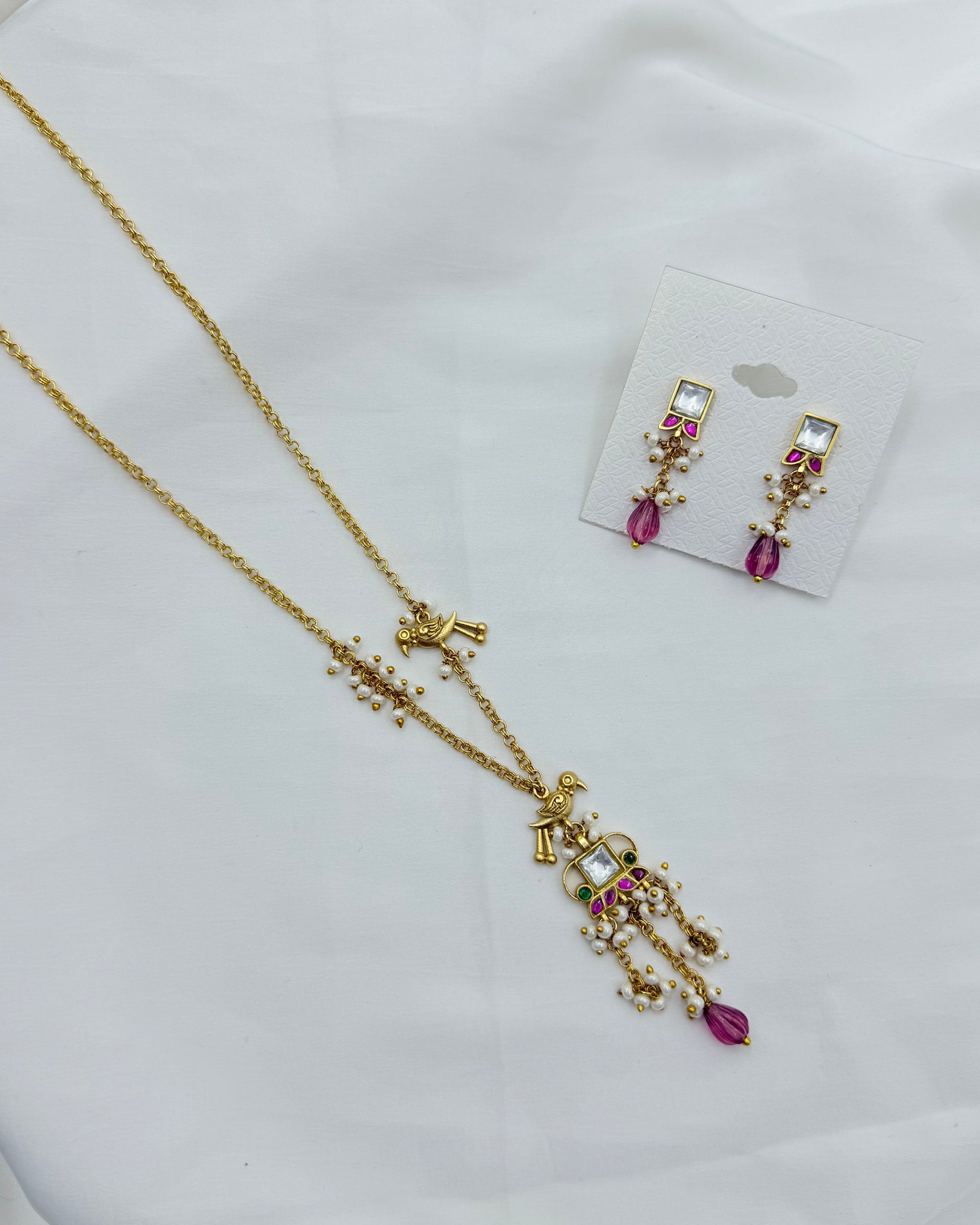 Gold-toned jewelry set featuring a necklace and matching earrings with pink gemstone accents and small clear stones, displayed on a white fabric background with the earrings on a small card.