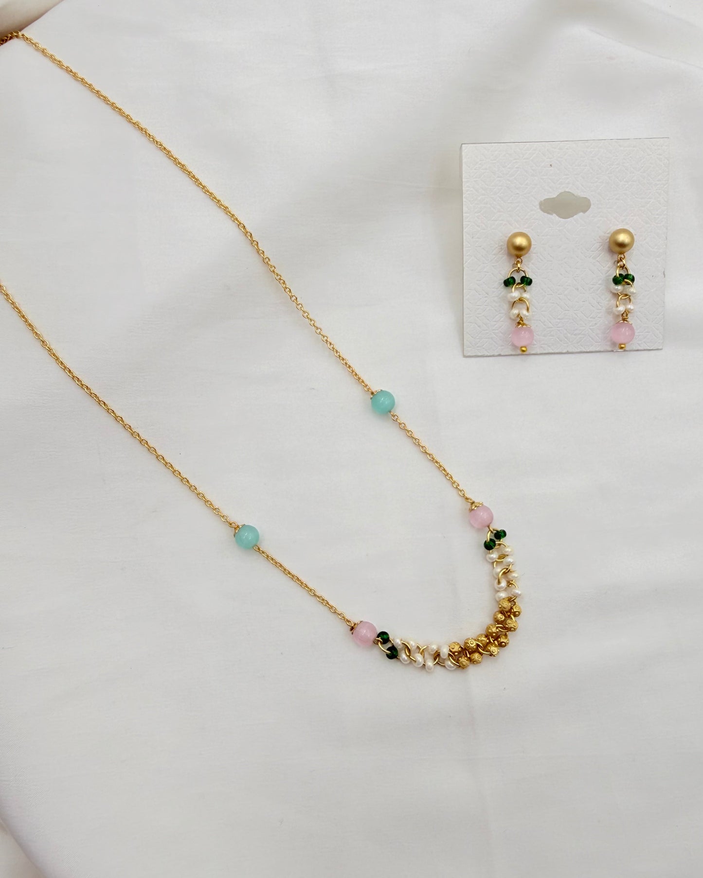 A gold necklace and matching drop earrings displayed on a white fabric surface, with the earrings attached to a small white card. The jewelry features gold beads, light blue beads, pale pink beads, and small dark green stones.
