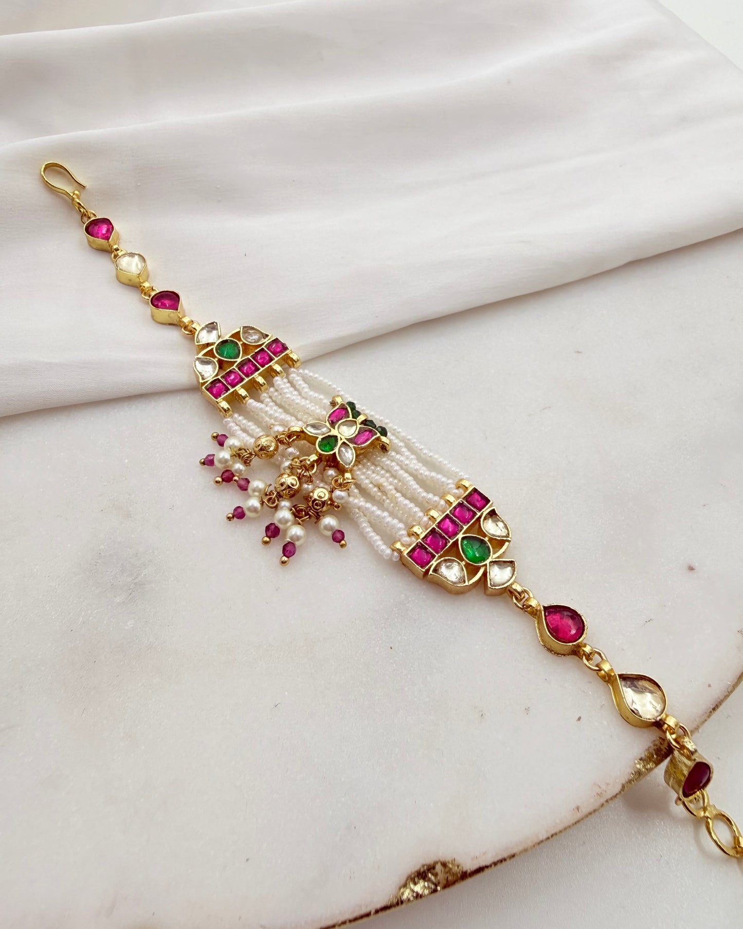 An ornate gold-plated traditional Indian style bracelet decorated with white faux pearls, pink and green stones, and small dangling bead accents, displayed on a draped white fabric background.