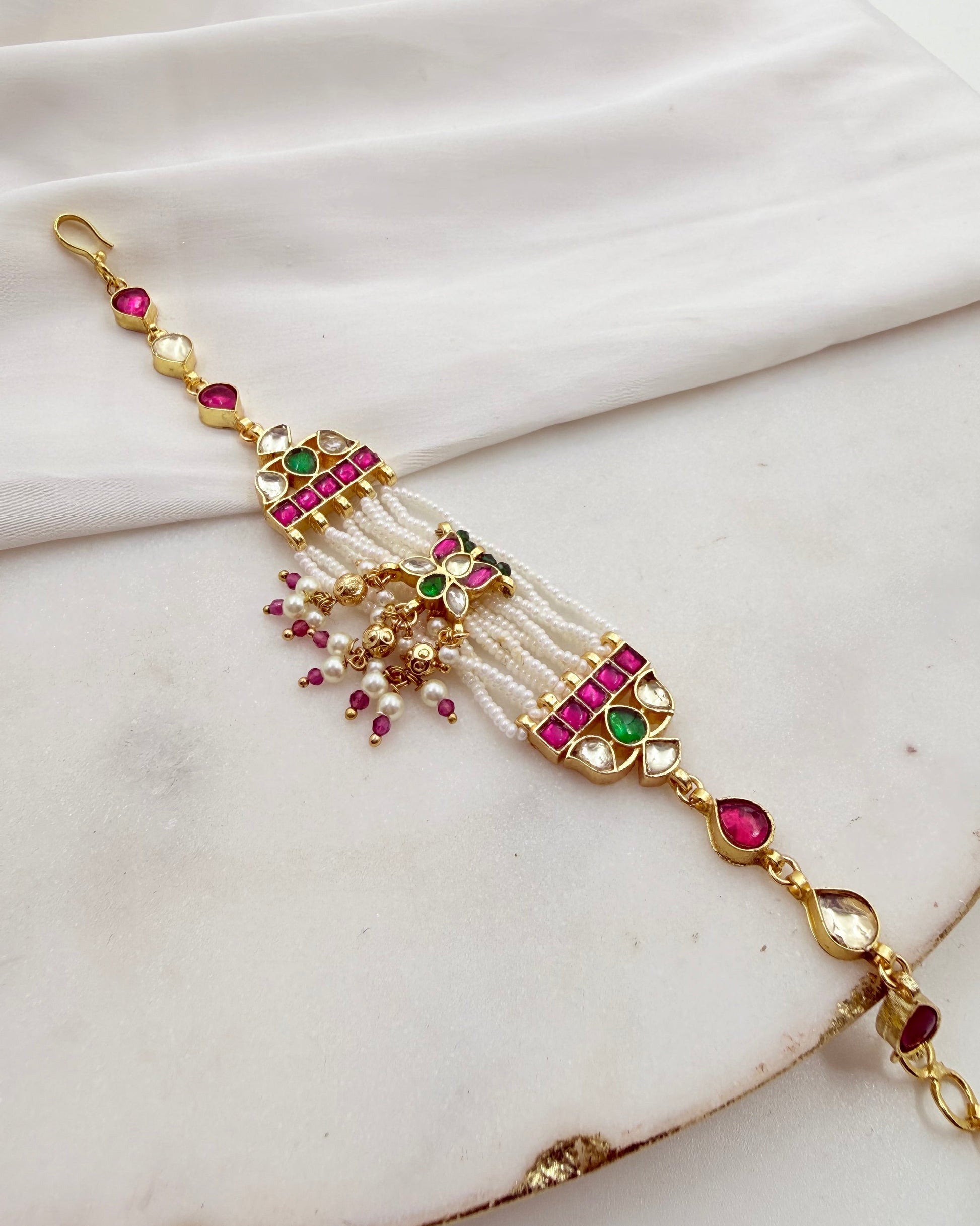 An ornate gold-plated traditional Indian style bracelet decorated with white faux pearls, pink and green stones, and small dangling bead accents, displayed on a draped white fabric background.