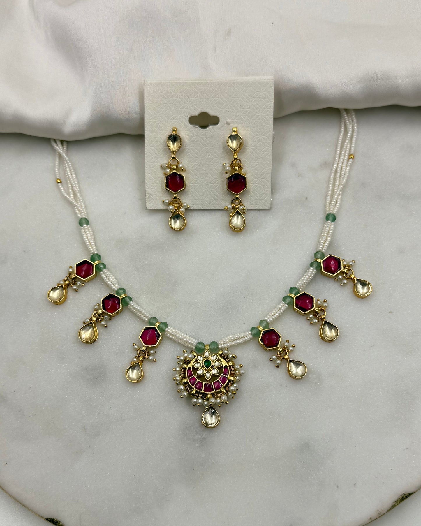 Gold and white jewelry set featuring a necklace and matching earrings with red and green stones, displayed on a white fabric and marble surface.