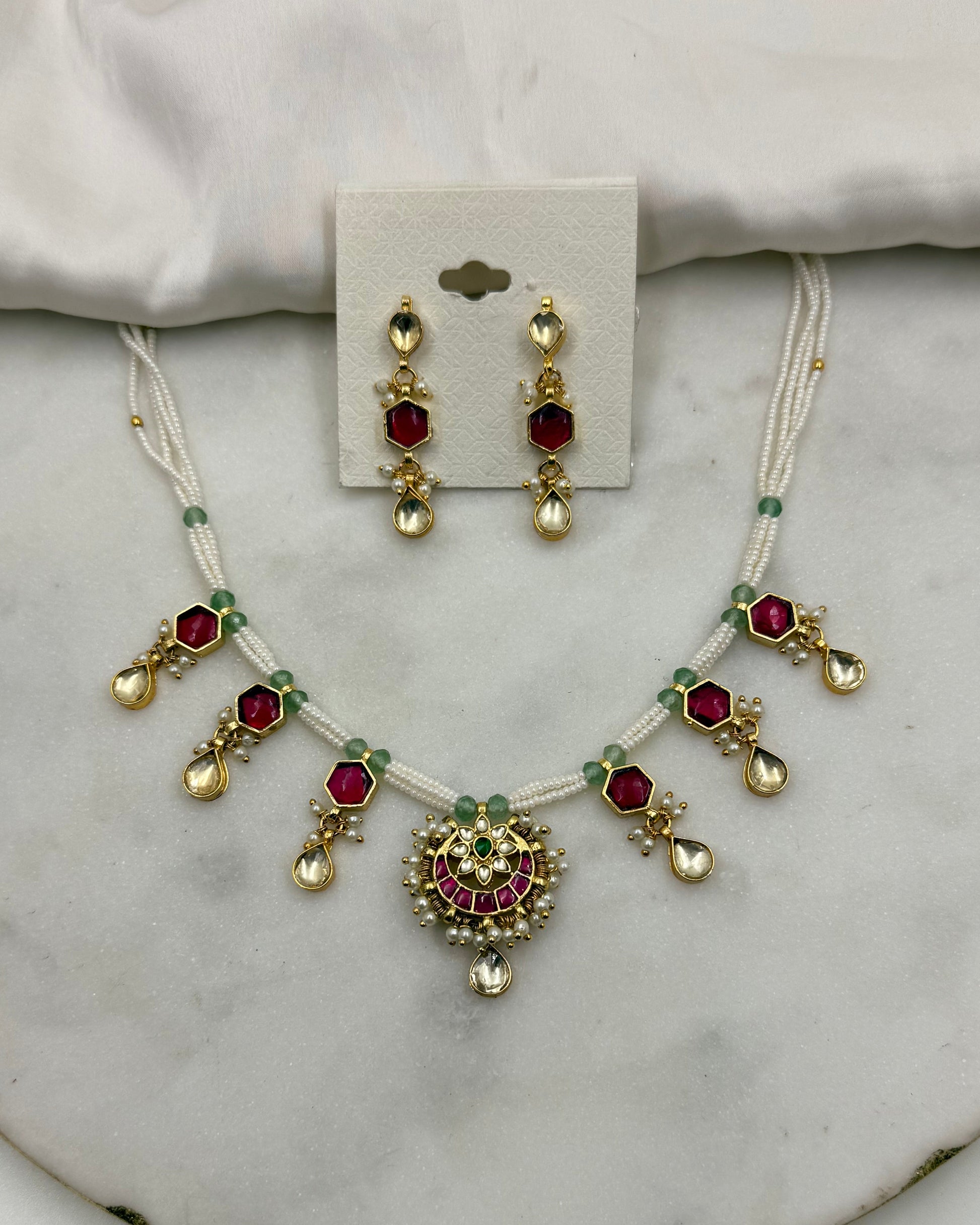 Gold and white jewelry set featuring a necklace and matching earrings with red and green stones, displayed on a white fabric and marble surface.