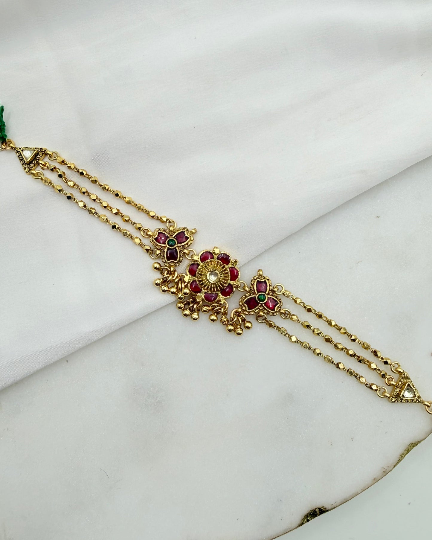 Gold-toned traditional Indian jewelry piece featuring a central floral motif with red and green stones, displayed on a white marble surface.